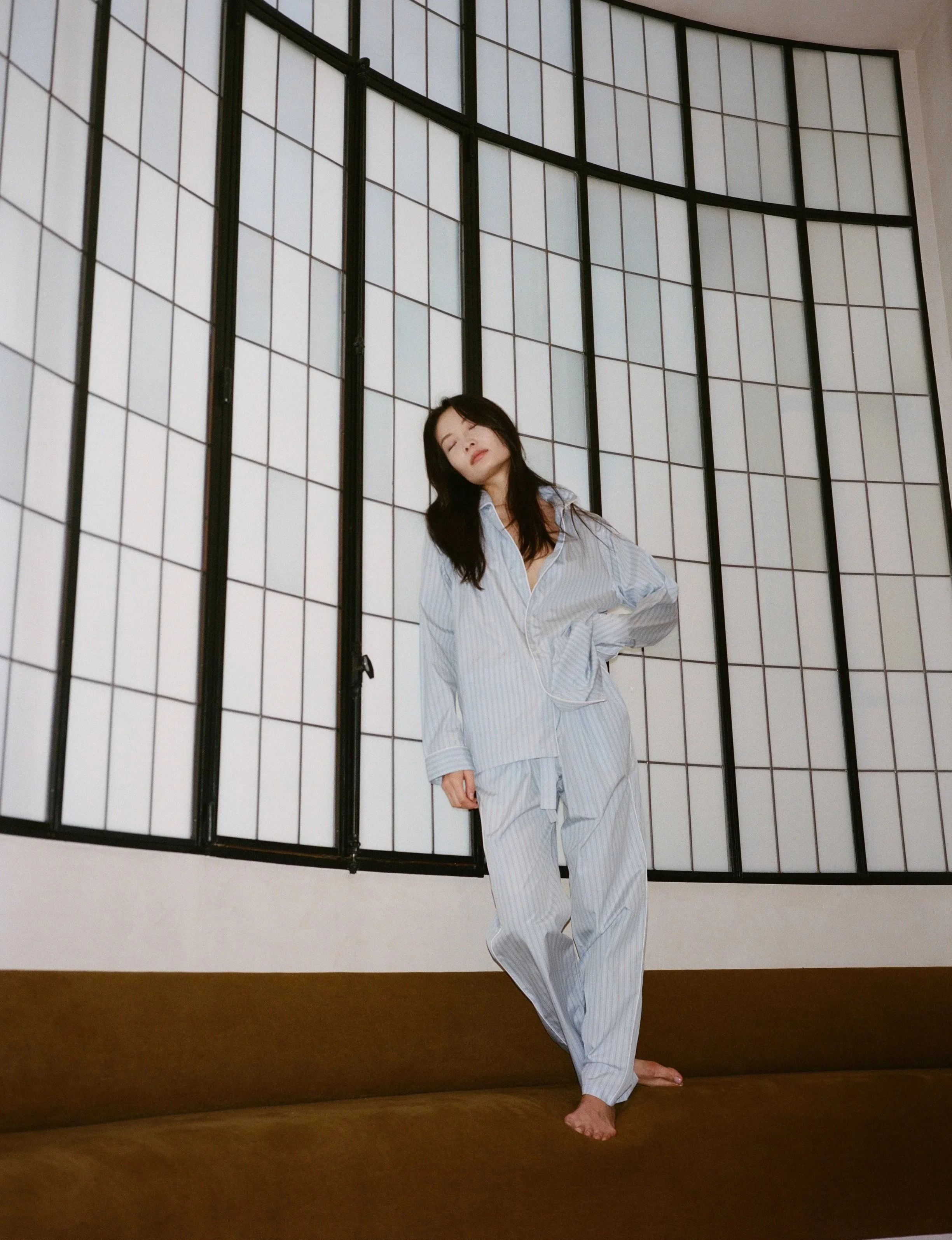 Eine Frau in Schlafanzug posiert vor einem weißen Raum mit schwarzen Gitterfenstern.
