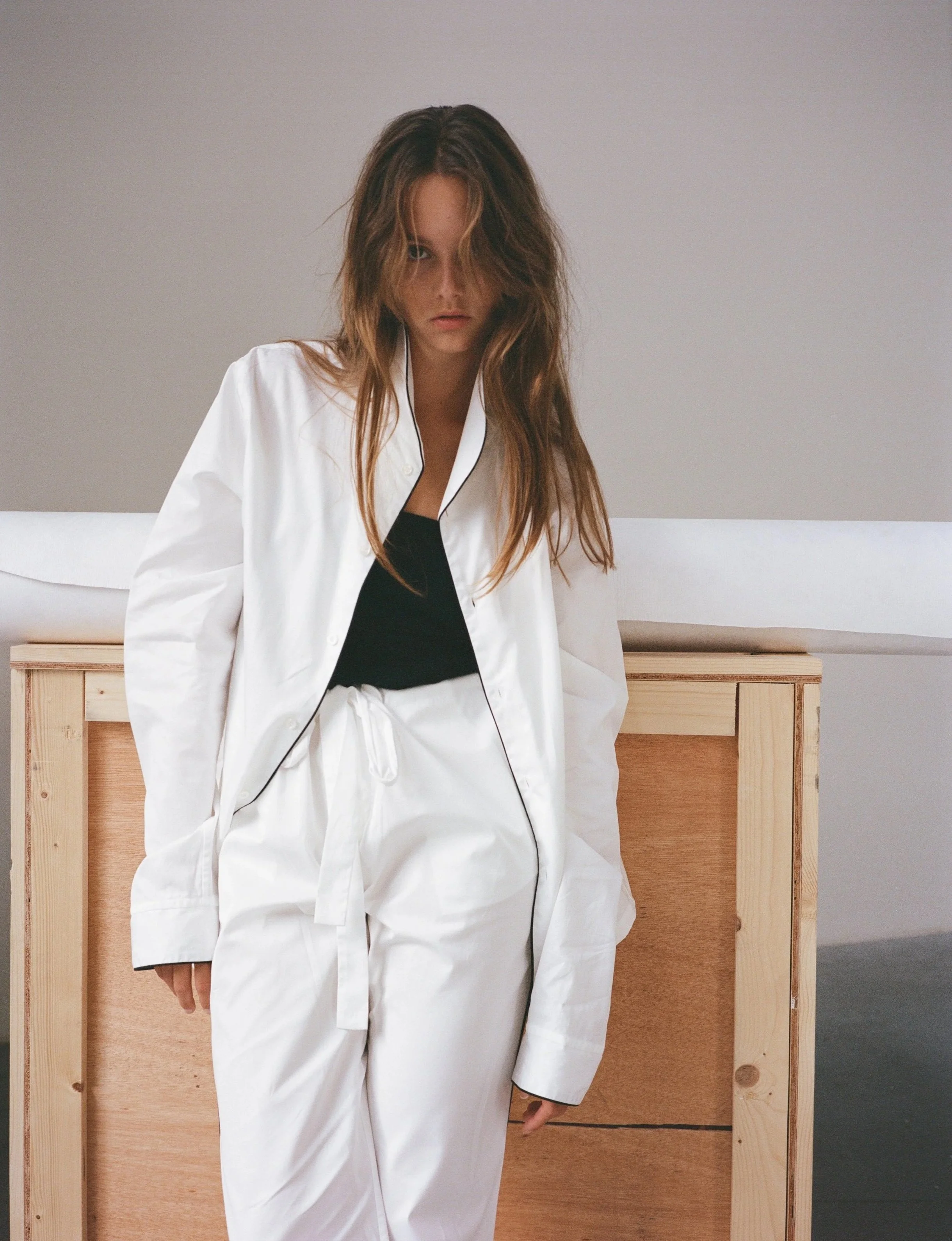 Eine junge Frau mit unordentlichen braunen Haaren trägt einen weißen Bademantel und ein schwarzes Top, steht vor einem Holzschrank in einem minimalistischen Raum.