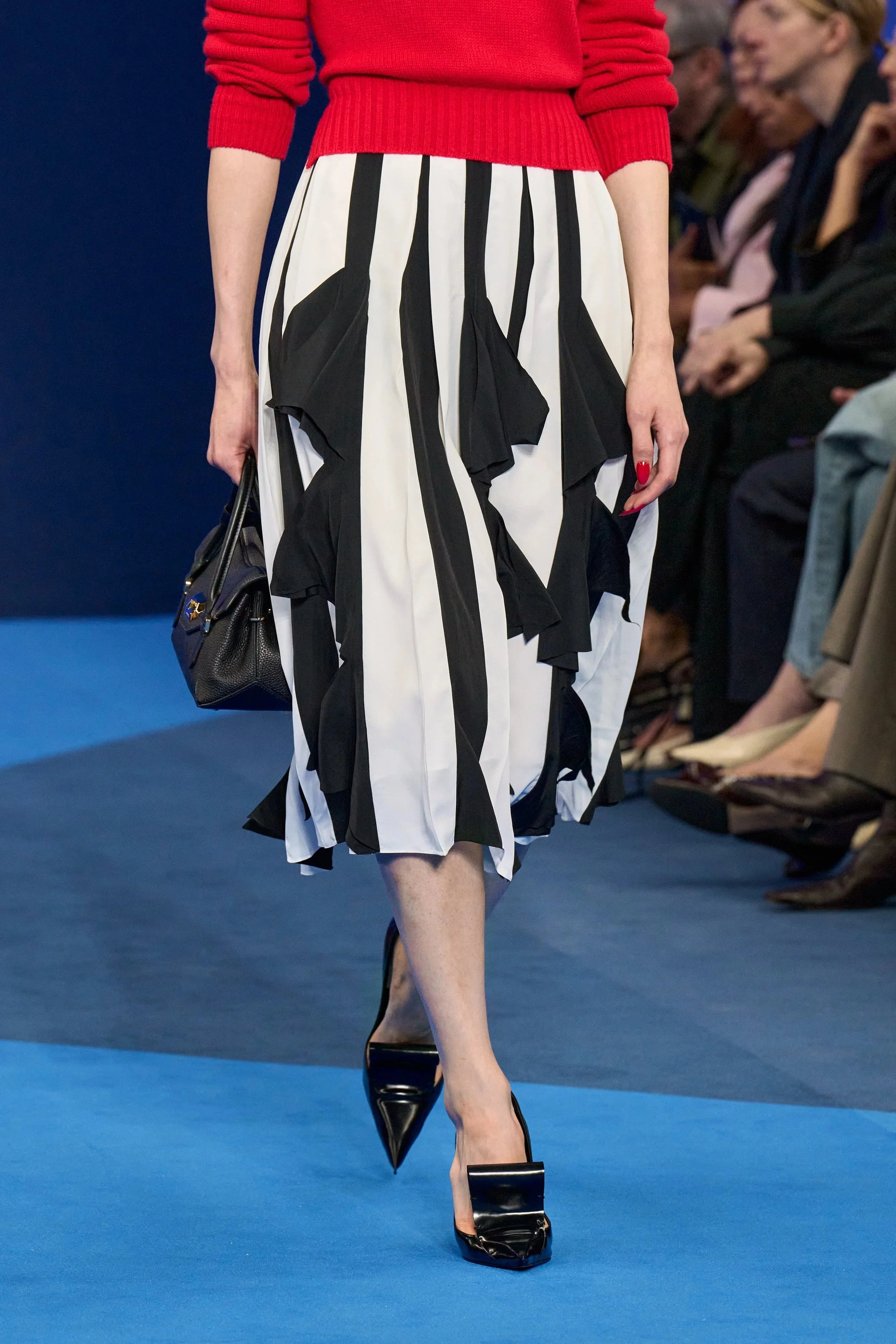 Eine Frau läuft auf dem Laufsteg, trägt ein rotes Oberteil, einen schwarz-weiß gestreiften und gepunkteten Rock, schwarze High Heels mit Metallelementen, und hält eine schwarze Handtasche.