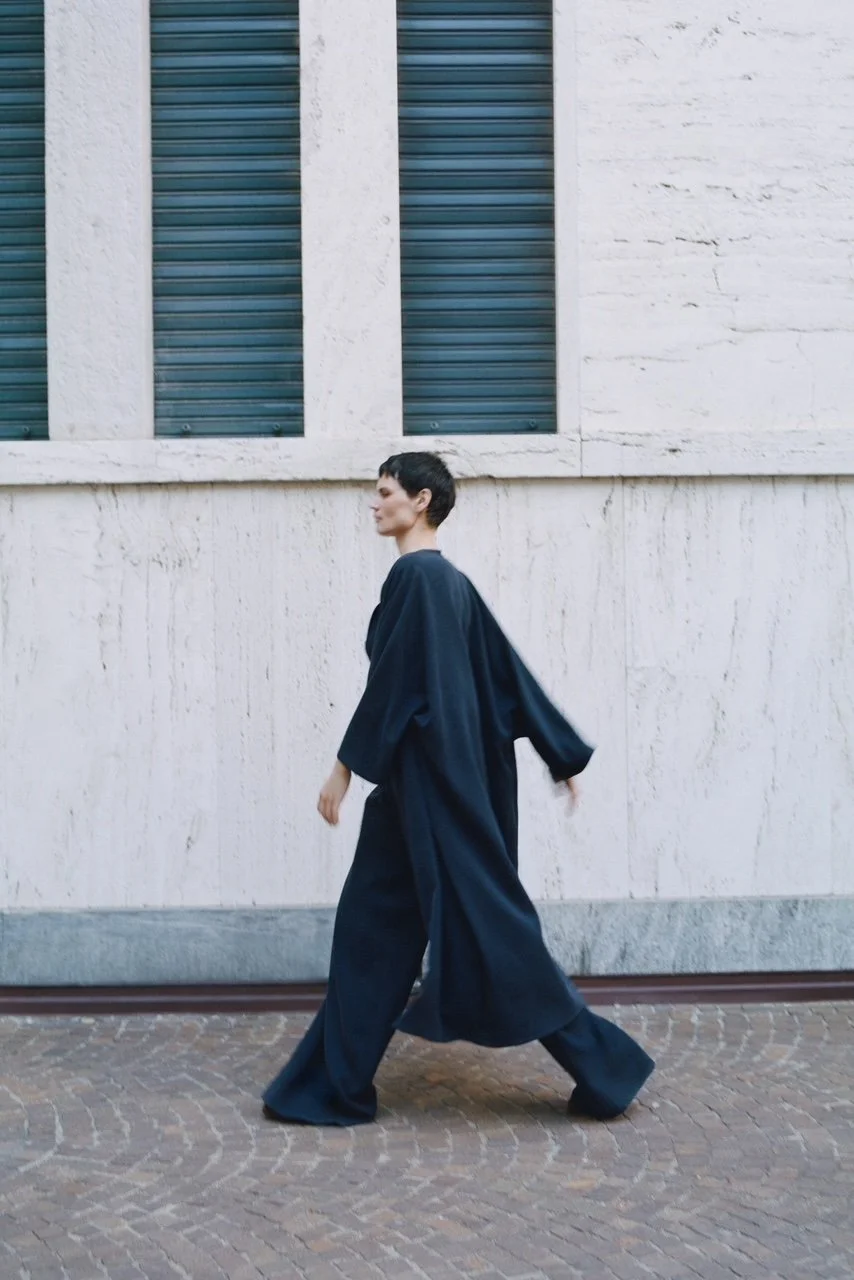 Eine Frau in dunkler, weiter Kleidung geht an einer weißen Wand entlang, im Hintergrund sind zwei Fenster mit blauen Rollläden.