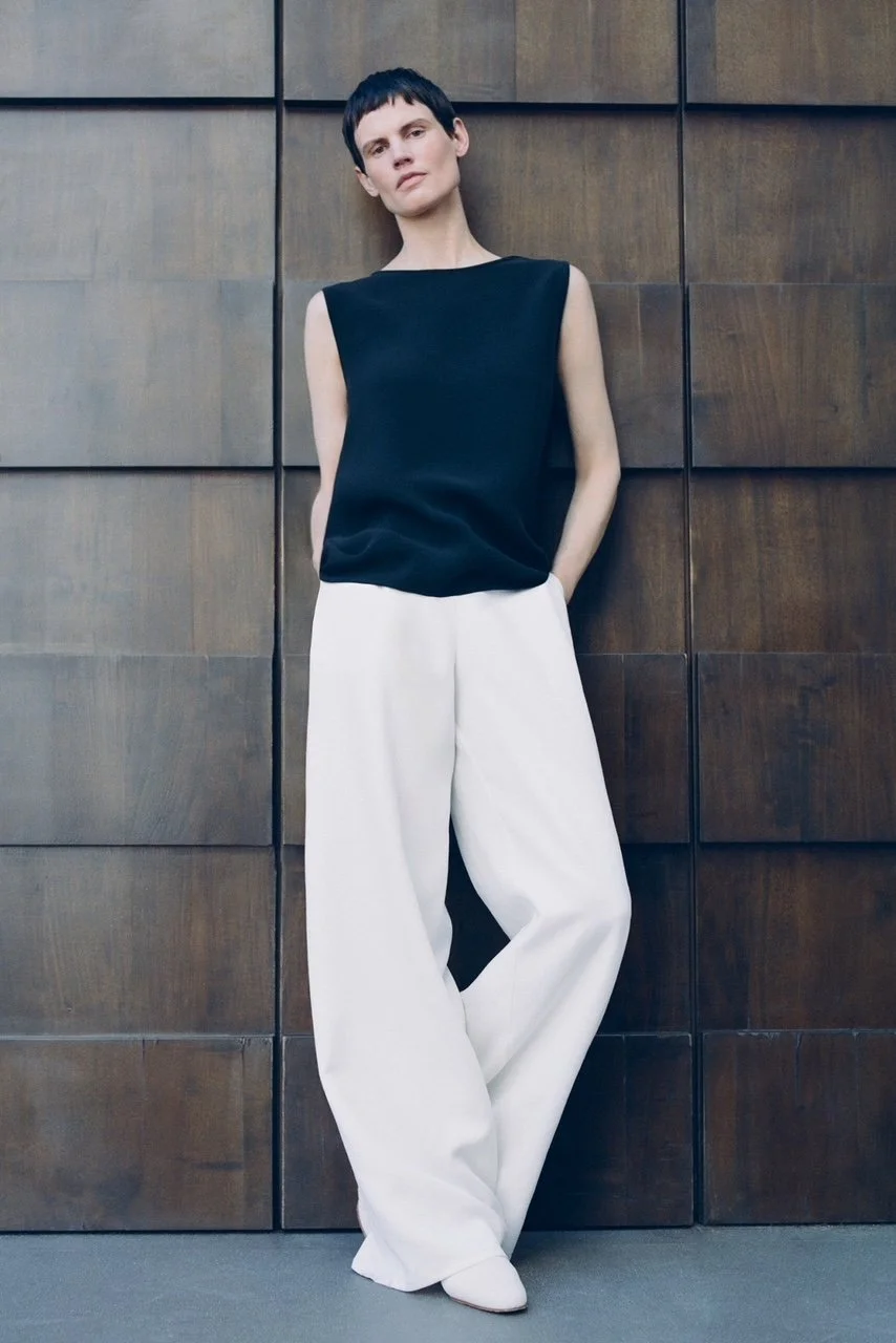 Frau mit kurzen schwarzen Haaren, schwarzes ärmelloses Top und weißen weiten Hosen, steht vor einer Holzwand.