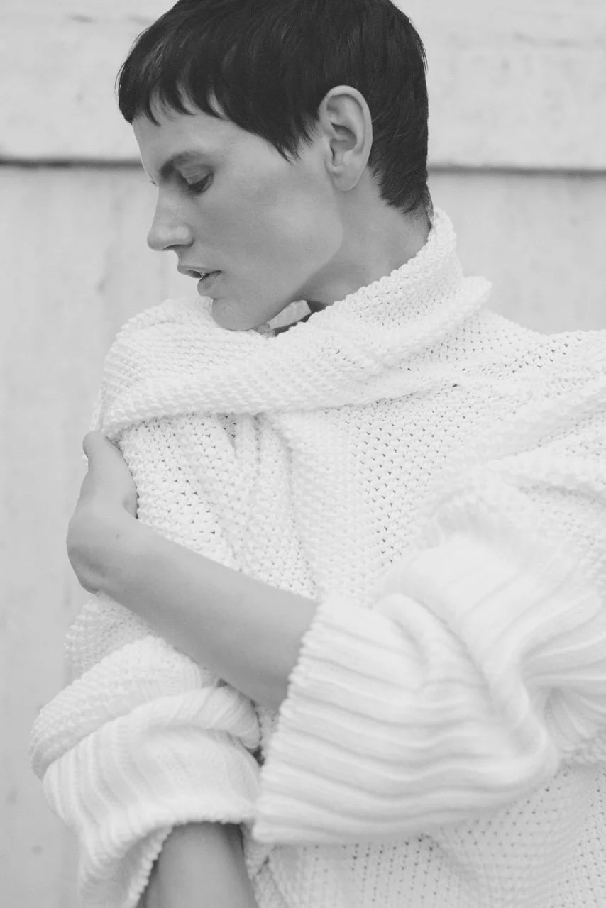 Schwarz-weiße Aufnahme einer Frau mit kurzen dunklen Haaren, die einen dick gestrickten weißen Schal trägt. Die Person ist nach links geneigt, scheint nachdenklich zu sein, und hält den Schal mit beiden Händen.