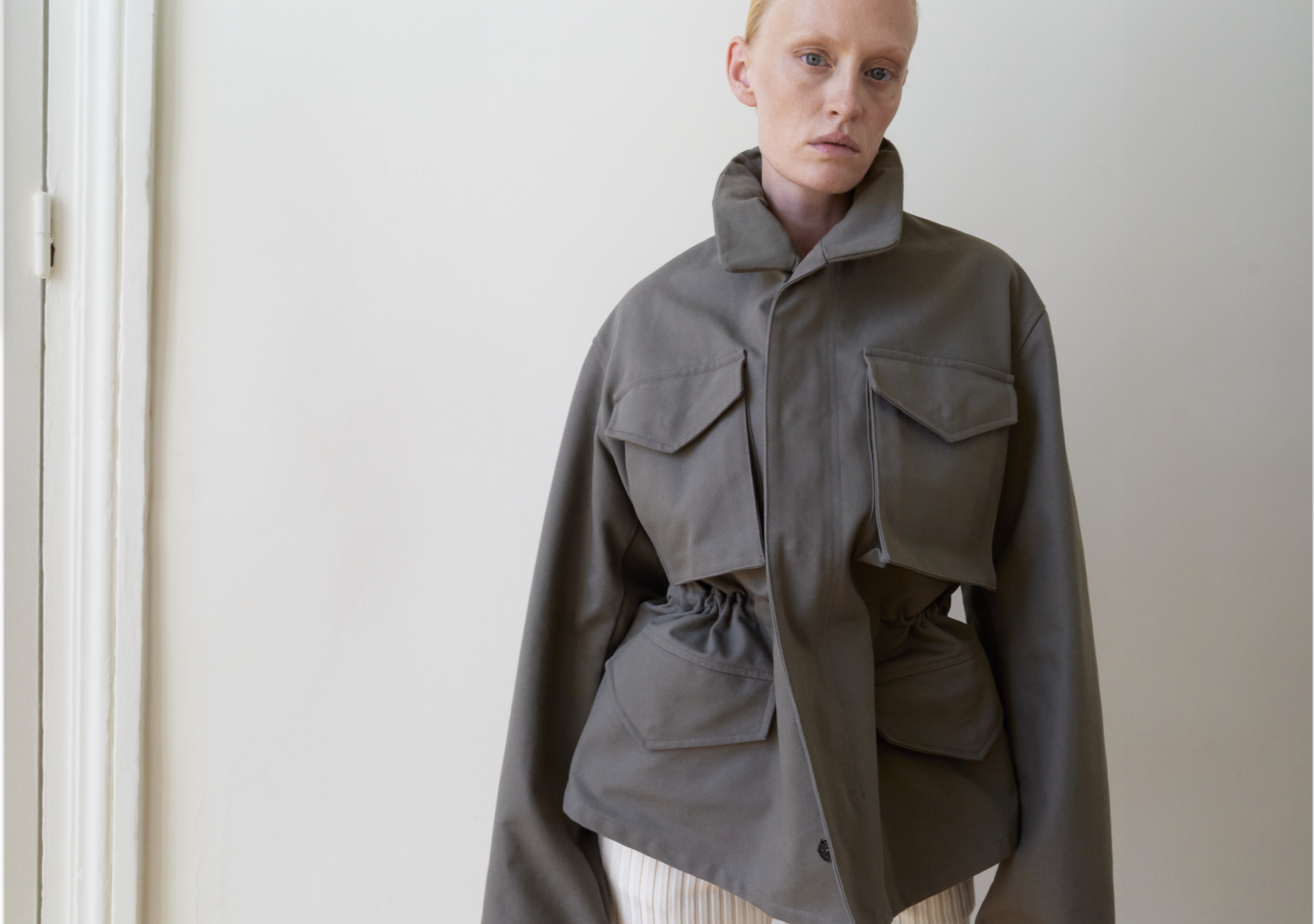 Frau in grauem Jacke mit großen Taschen, steht vor weißer Wand.