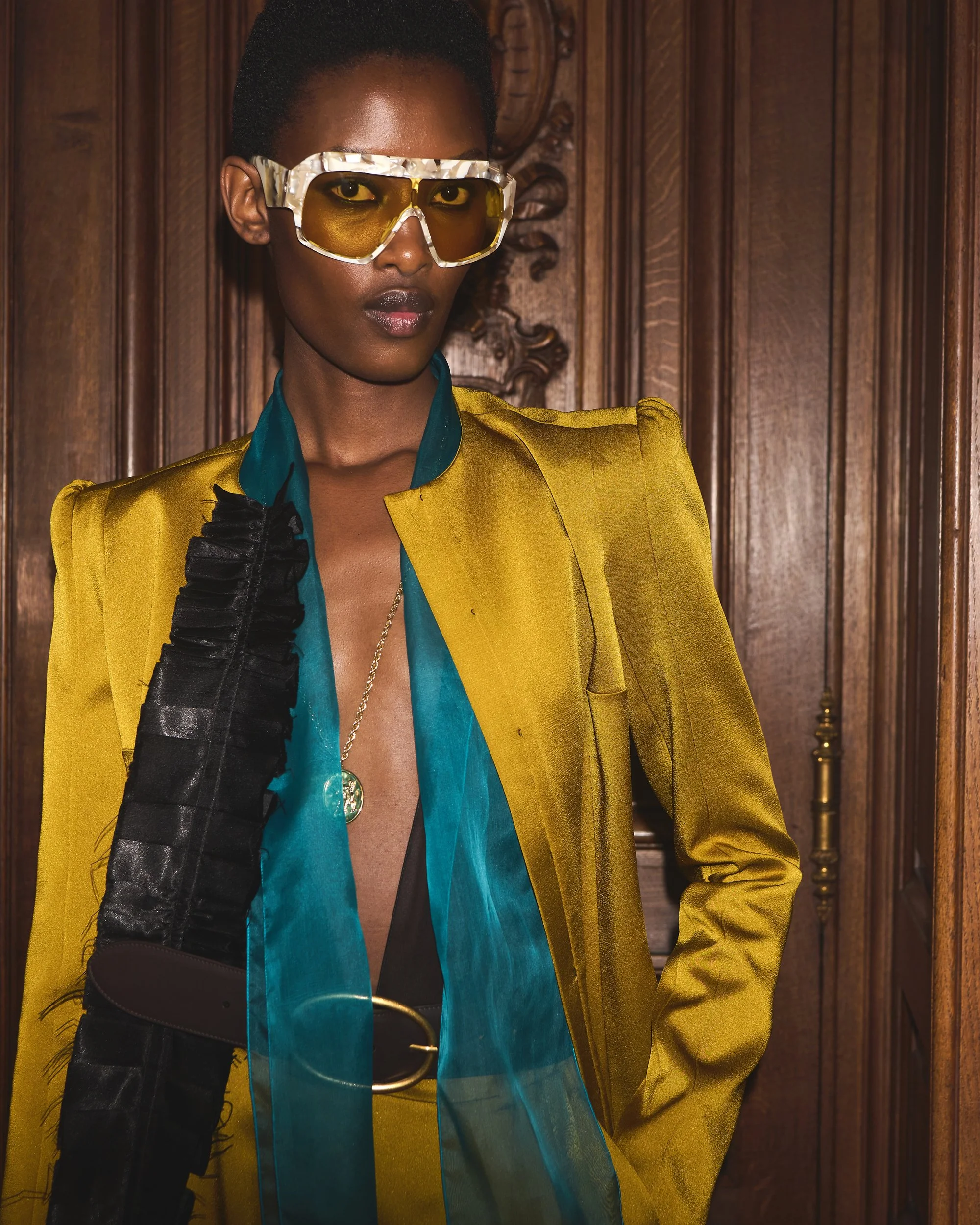 Frau trägt eine gelbe Jacke, blaue Bluse, eine schwarze Schultertasche und trägt gelbe Sonnenbrille vor einer Holzvertäfelung.
