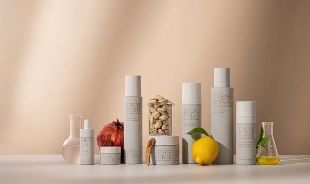 Verschiedene Pflegeprodukte in weißen Flaschen, sowie eine Granatapfel, Zitrone, einige Pistazien, ein Blatt und ein kleines Fläschchen mit Öl auf einer weißen Oberfläche.