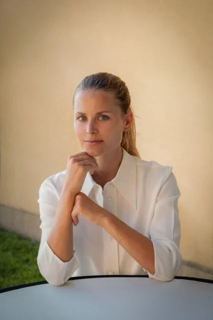 Junge Frau mit blonden Haaren in weißem Hemd sitzt an einem Tisch vor einem hellen Hintergrund.
