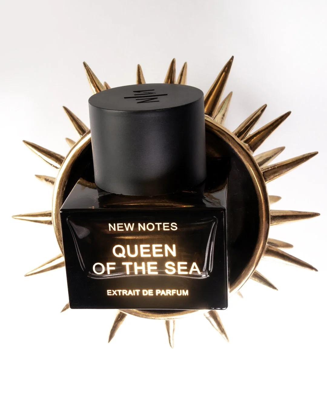 Schwarbe Parfümflasche 'Queen of the Sea' auf einer goldenen Spikes-Merch, vor weißem Hintergrund.
