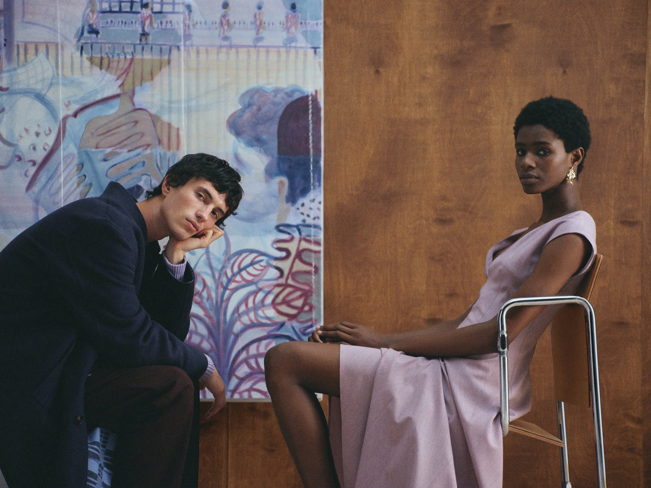 Ein junger Mann und eine junge Frau sitzen in einem Raum, der mit Holzpanelen verkleidet ist. Der Mann sitzt auf einem Stuhl auf dem Boden und stützt den Kopf mit der Hand, während er in die Kamera schaut. Das Mädchen sitzt auf einem Stuhl, leicht nach vorne gebeugt, mit einem ernsten Blick zur Kamera. Im Hintergrund ist eine farbenfrohe Wandmalerei zu sehen.