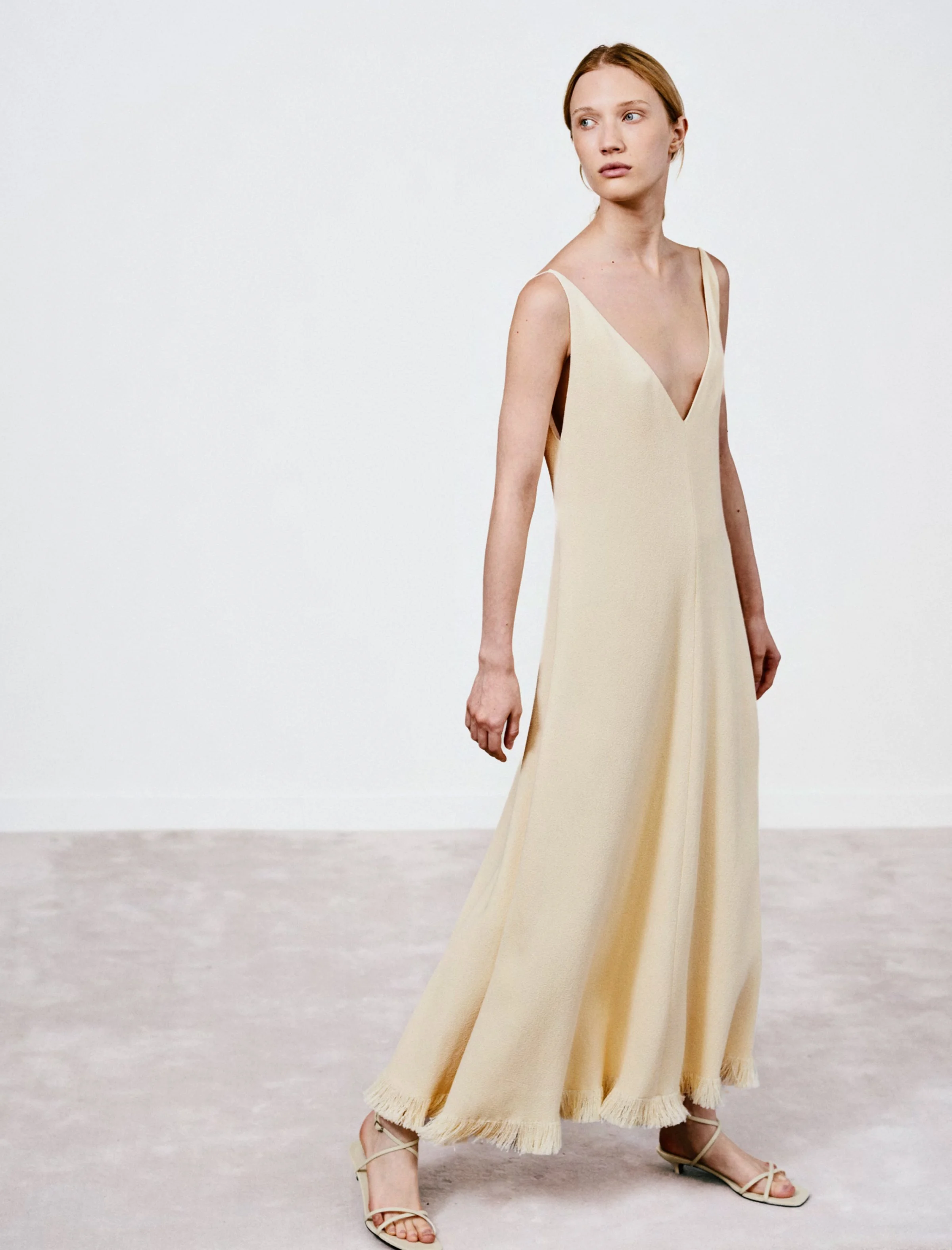 Eine junge Frau trägt ein langes, cremefarbenes Kleid mit Fransen am Saum, offene Sandalen, steht auf einem glatten Boden vor einer weißen Wand.