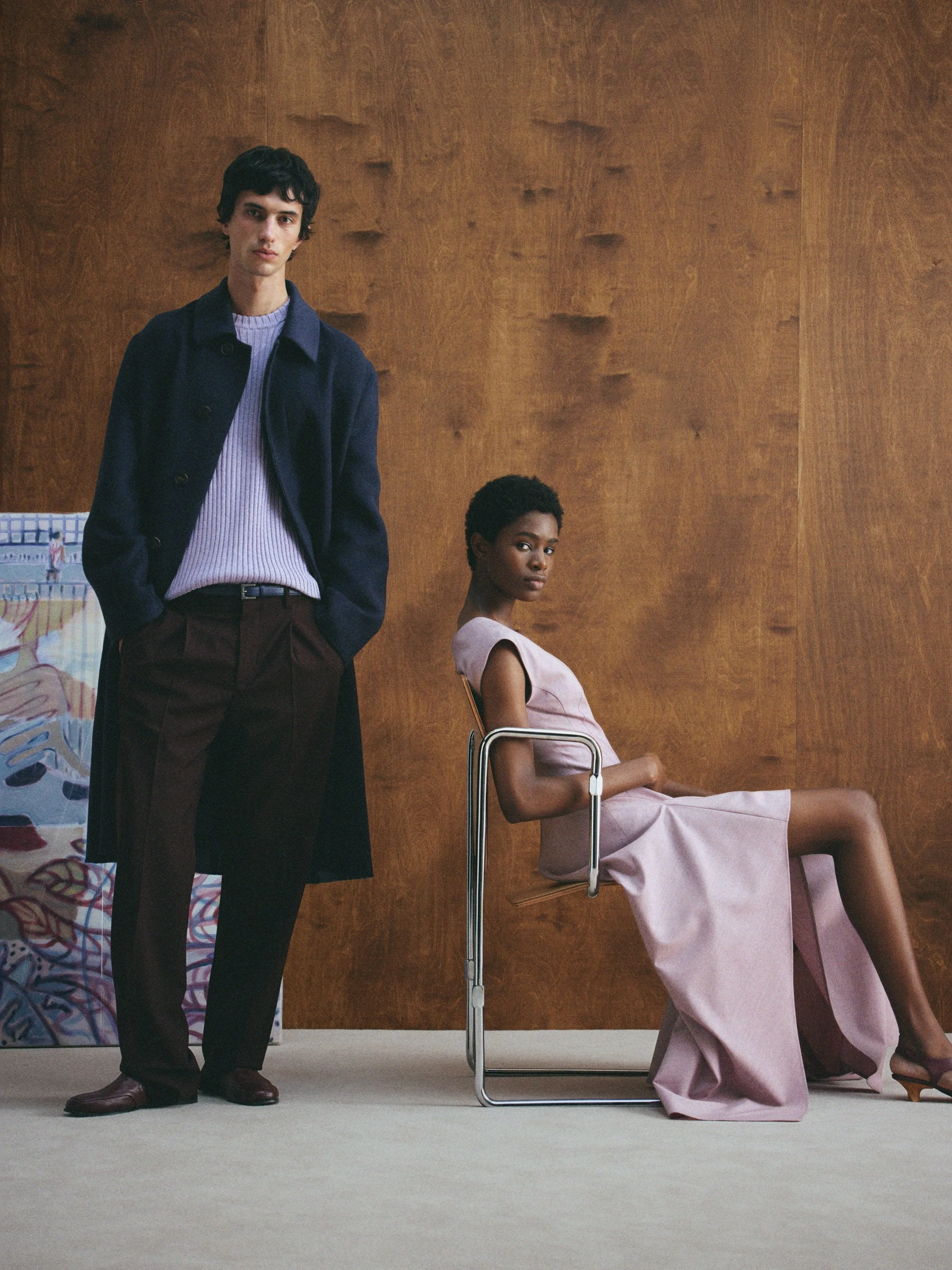 Ein Mann steht links, trägt einen dunklen Mantel und braune Hosen, eine Frau sitzt rechts in einem Stuhl, trägt ein rosa Kleid, beide vor einer Holzwand.