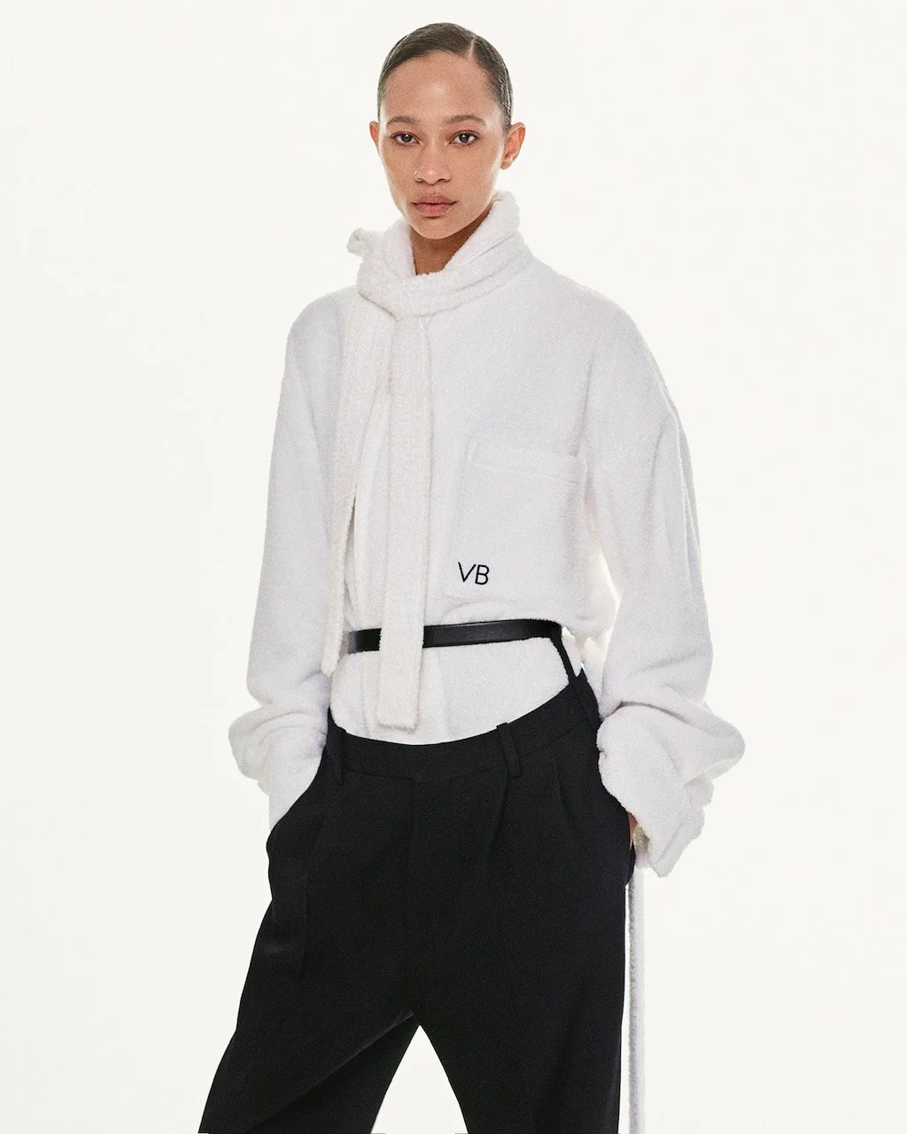 Junge Frau in weißer Jacke und schwarzen Hosen, steht vor weißem Hintergrund.