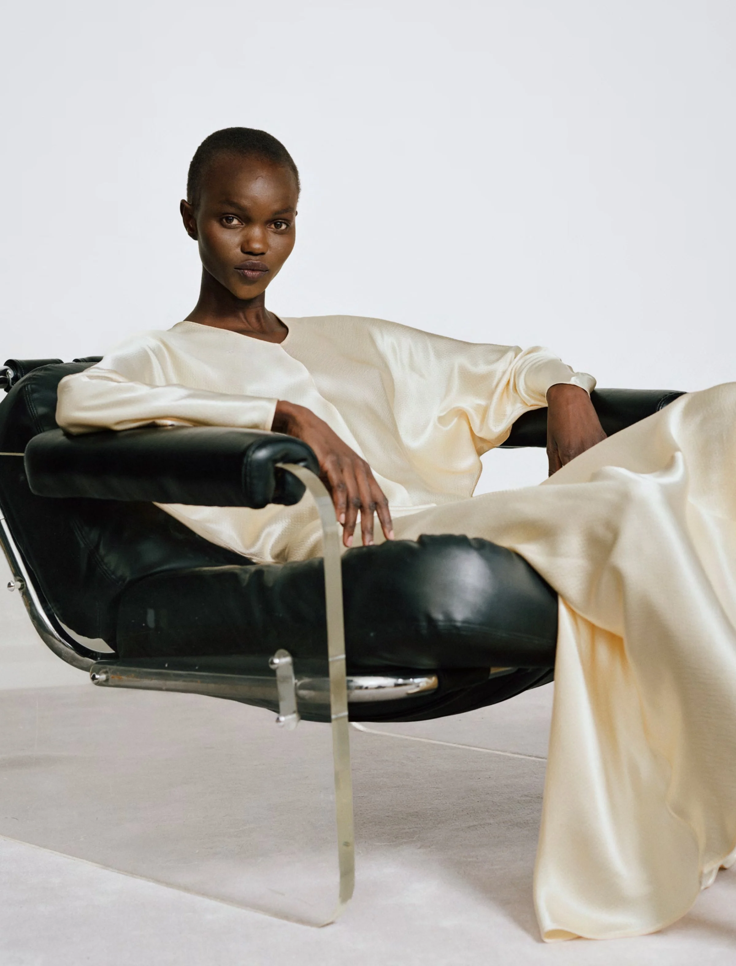 Eine schwarze Frau sitzt entspannt auf einem modernen schwarzen Lederstuhl in einem weißen Raum, trägt ein langes, cremefarbenes Kleid.
