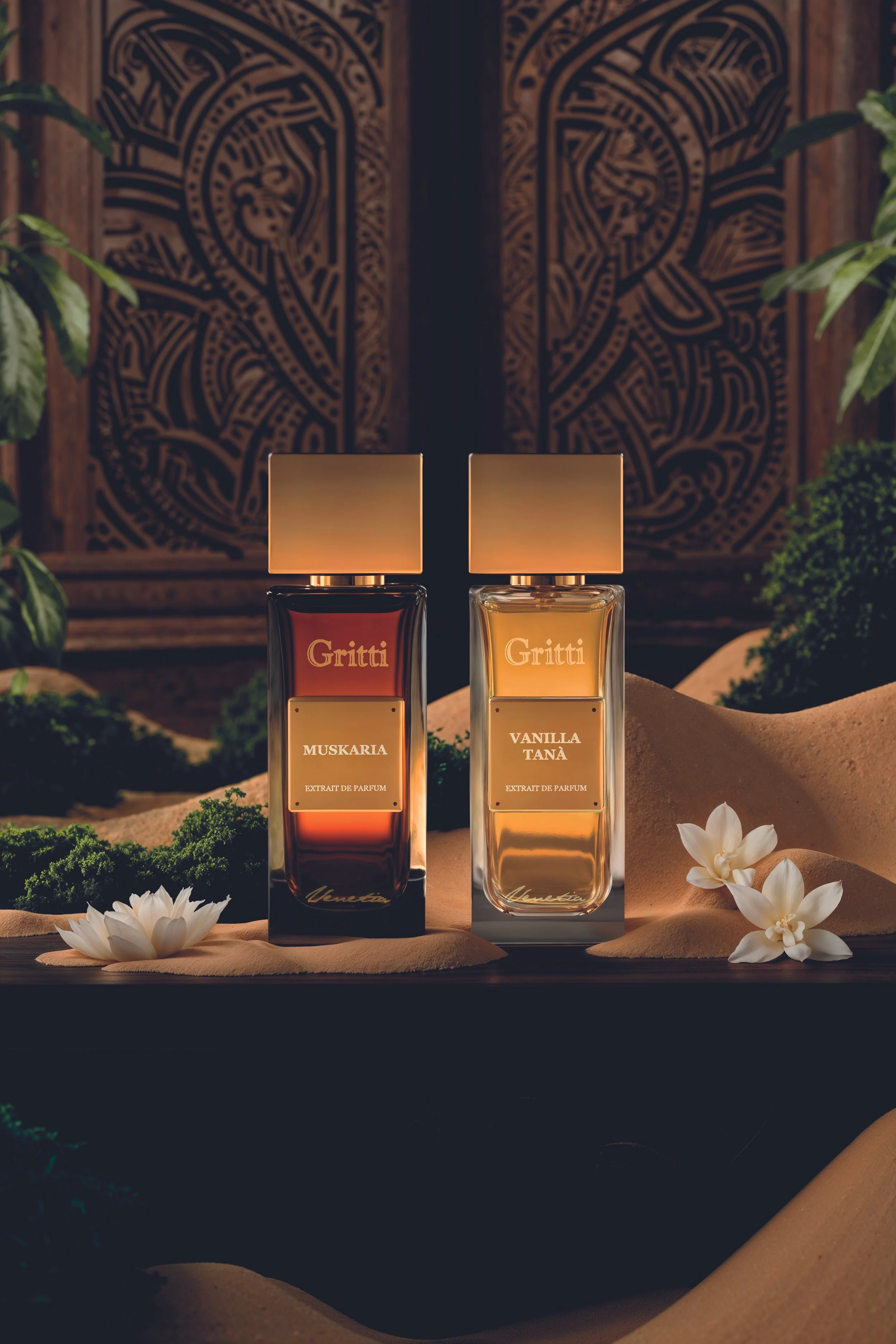 Zwei Parfümflaschen auf einer schwarzen Oberfläche, umgeben von weißen Blumen, mit einer sandfarbenen Stoffunterlage im Hintergrund, vor einem kunstvollen Holzschnitzereimotiv.