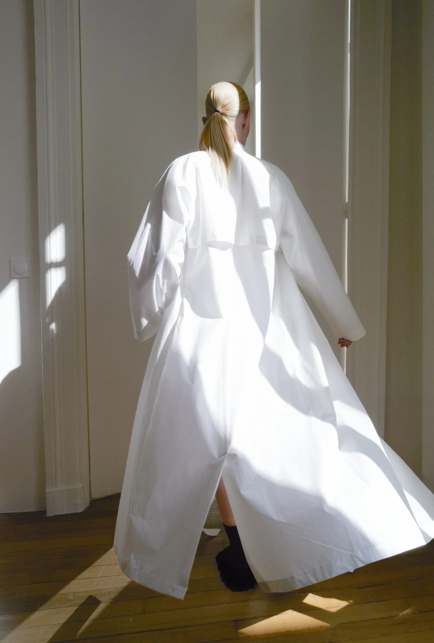 Person in weißem Kleid oder Kleidungsstück, die einen Raum mit natürlichem Licht betreten, Schatten an der Wand.