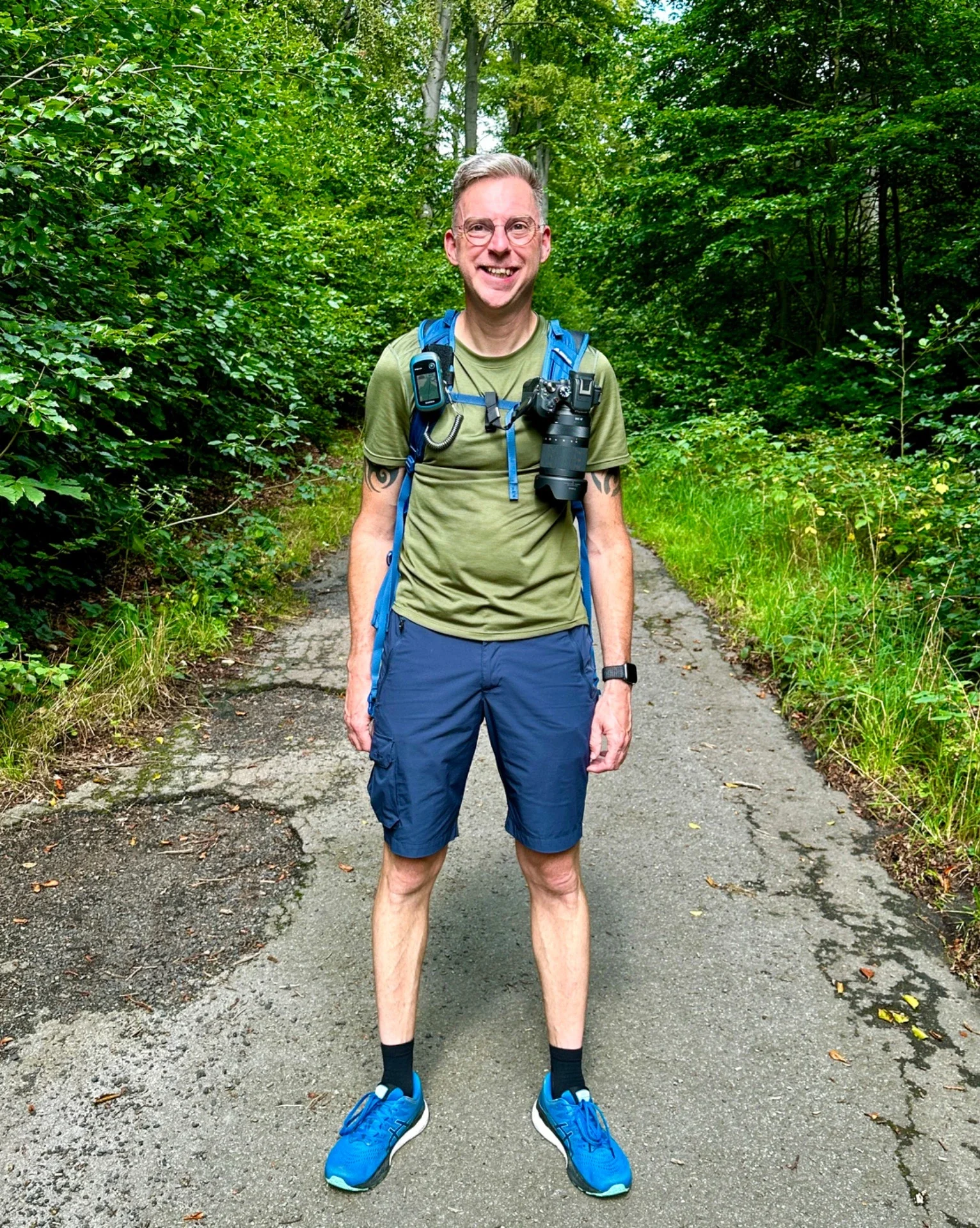 Man in casual outdoor clothing standing on a forest trail, smiling, with camera gear around neck and backpack.