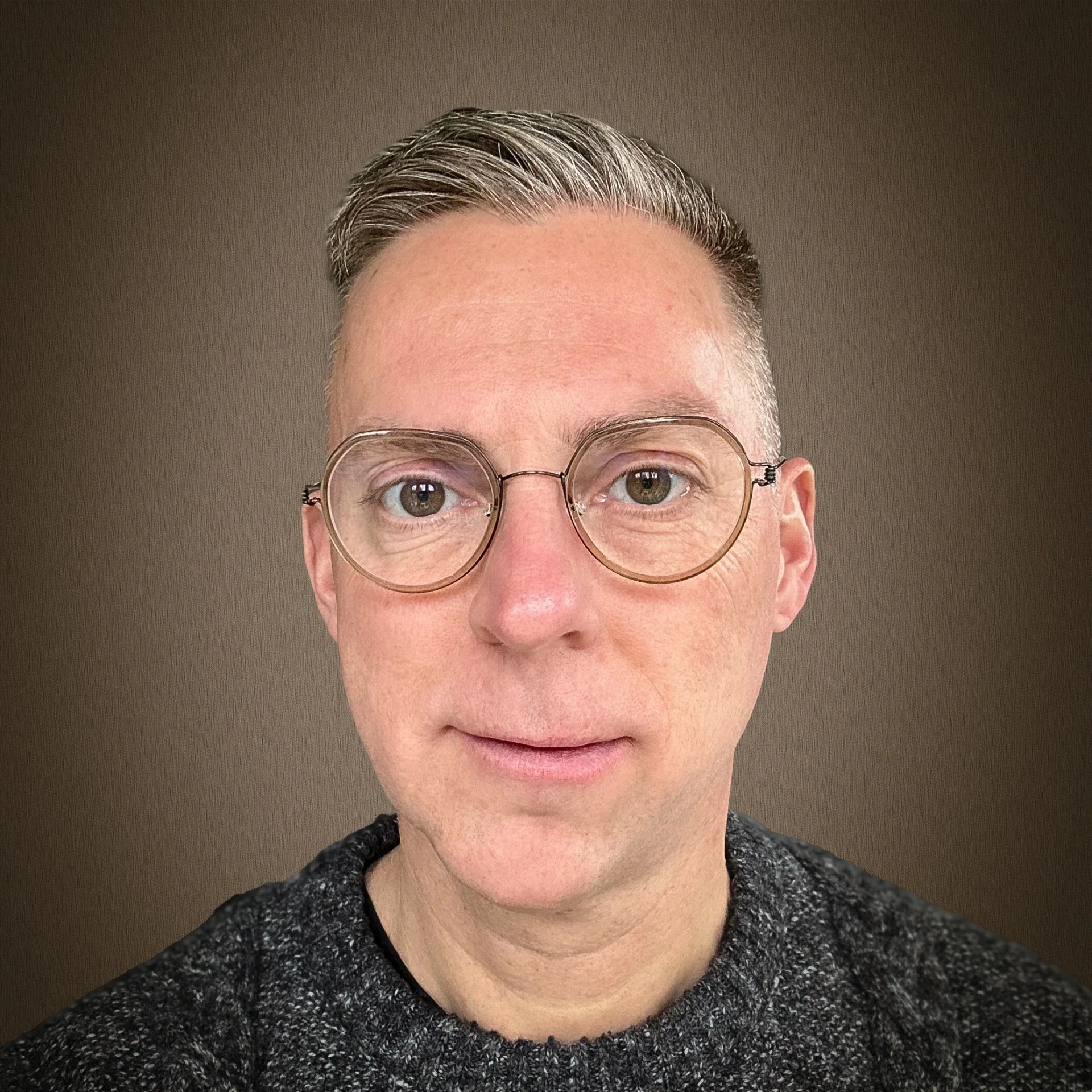 Portrait of a man with short, gray hair, wearing round glasses and a dark gray sweater, against a plain brown background.