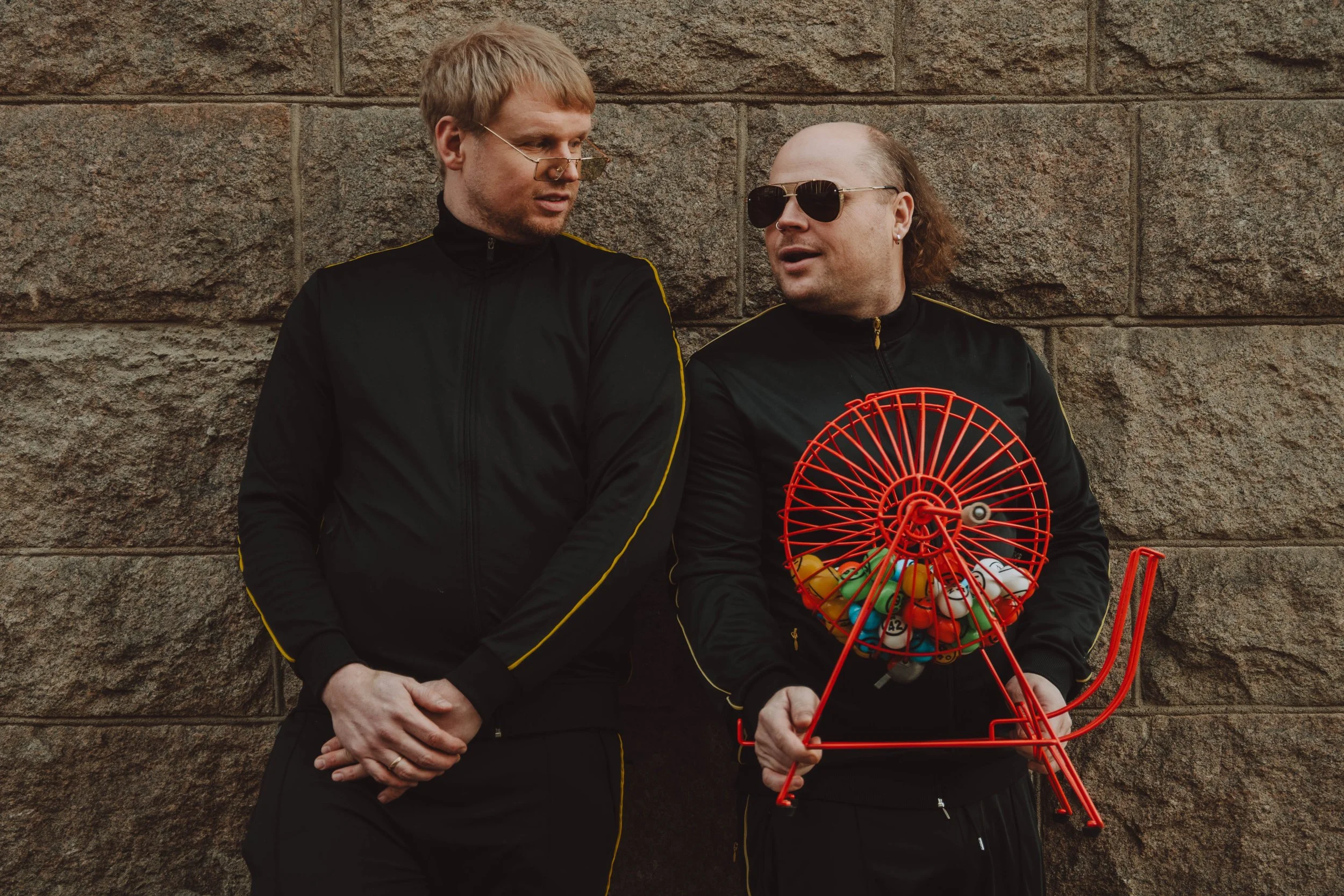 To menn står mot en steinvegg, en holder en liten rød radiator med baller, den andre snakker. Begge har mørke treningsdresser med gule stripper og solbriller.