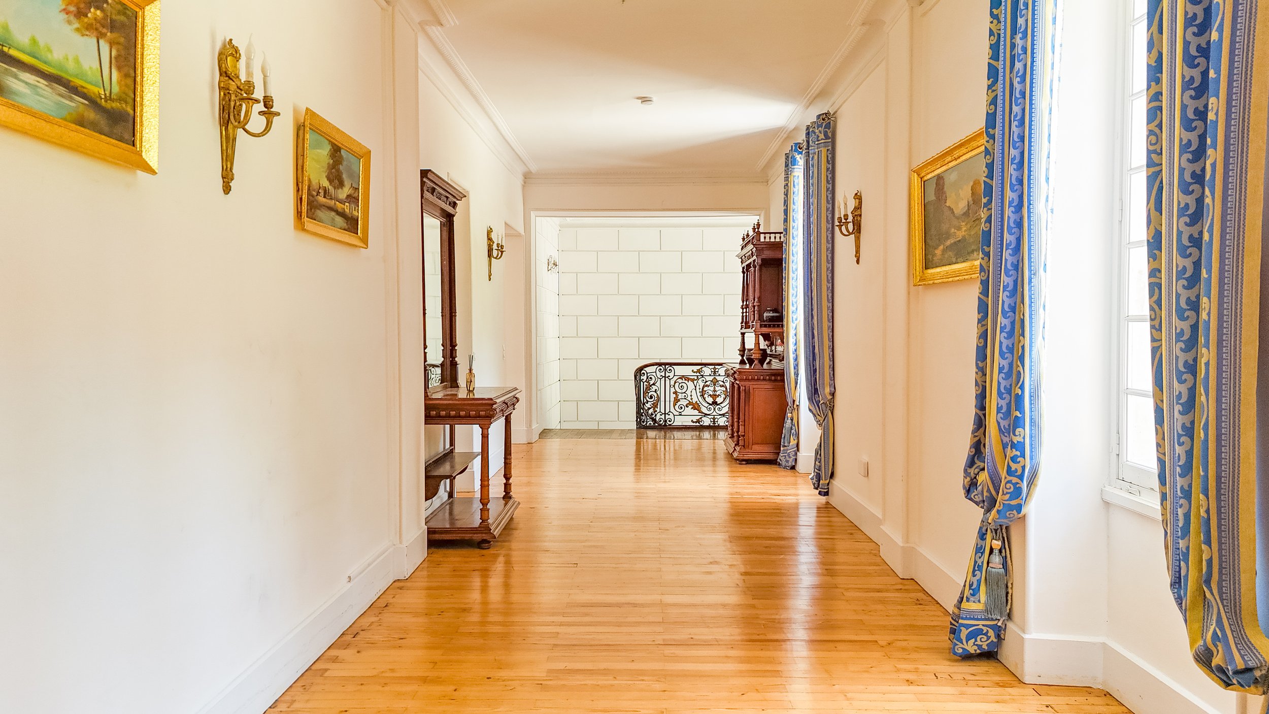 Intérieur d'une pièce avec des murs blancs, des cadres avec des peintures, des rideaux bleus avec des motifs dorés, et un parquet en bois.