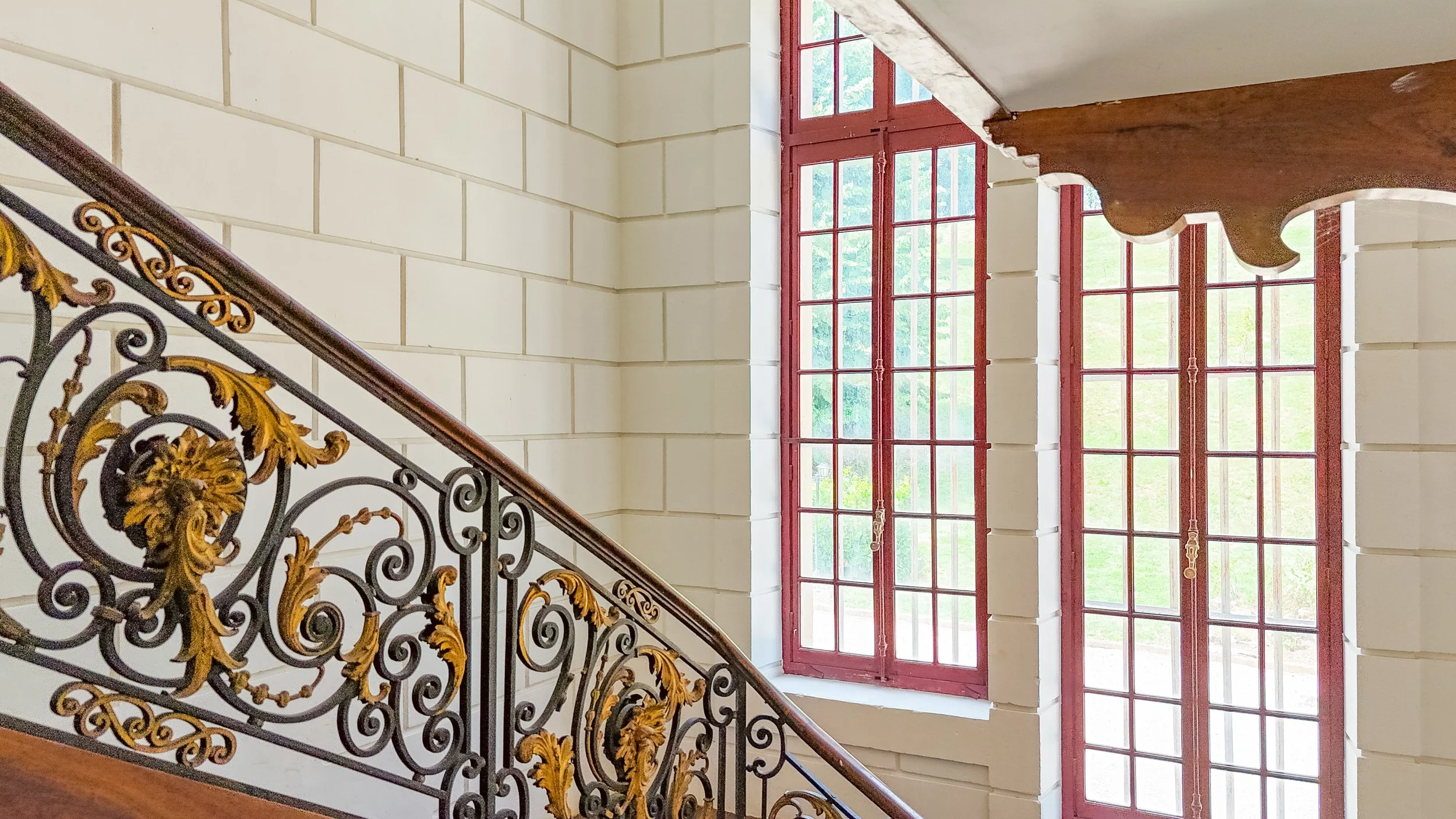 Escalier avec rampe en métal noir et or, murs en pierre claire, grandes fenêtres à cadre rouge laissant passer la lumière naturelle.
