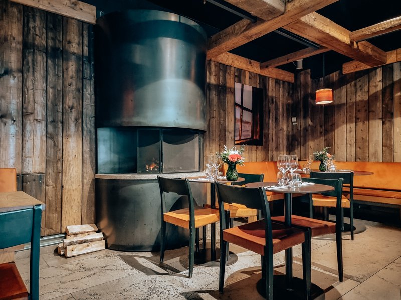 A cozy restaurant corner with a wood-paneled wall, a fireplace, and a table set with wine glasses and floral arrangements.