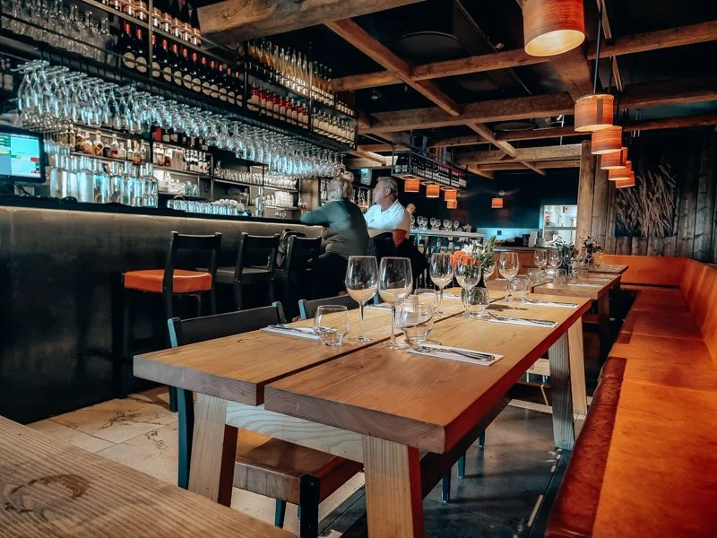 A stylish restaurant interior with a bar area, wooden dining tables set with glasses, plates, and napkins, and cozy orange bench seating along the wall. Decorative shelves are filled with glassware and bottles, and pendant lights hang from the ceilin