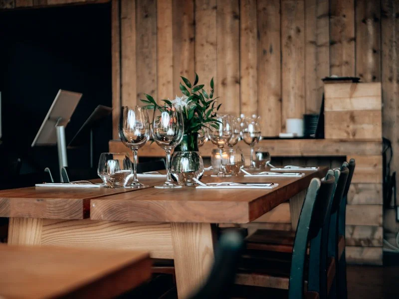 Elegant dining table set with wine glasses, water glasses, white napkins, and cutlery, with a potted plant centerpiece in a rustic wooden interior.