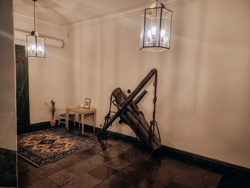 A furnished corner of a hallway with two hanging light fixtures, a small table with a picture frame and dried plant in a vase, a decorative rug, and a large vintage wooden camera on a tripod leaning against the wall.