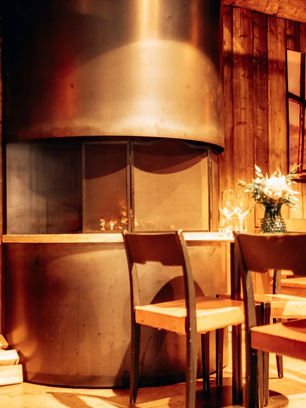 Indoor cozy setting with a stainless steel fireplace, wooden chairs, and a vase with white flowers on a wooden table.