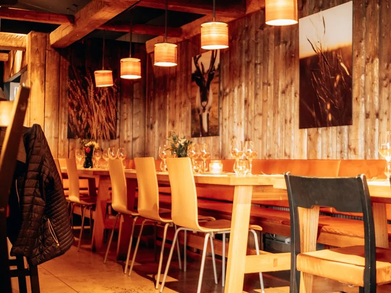Cozy restaurant interior with wooden walls, hanging pendant lights, and artwork of wildlife, set for dining with glasses and flowers on the tables.