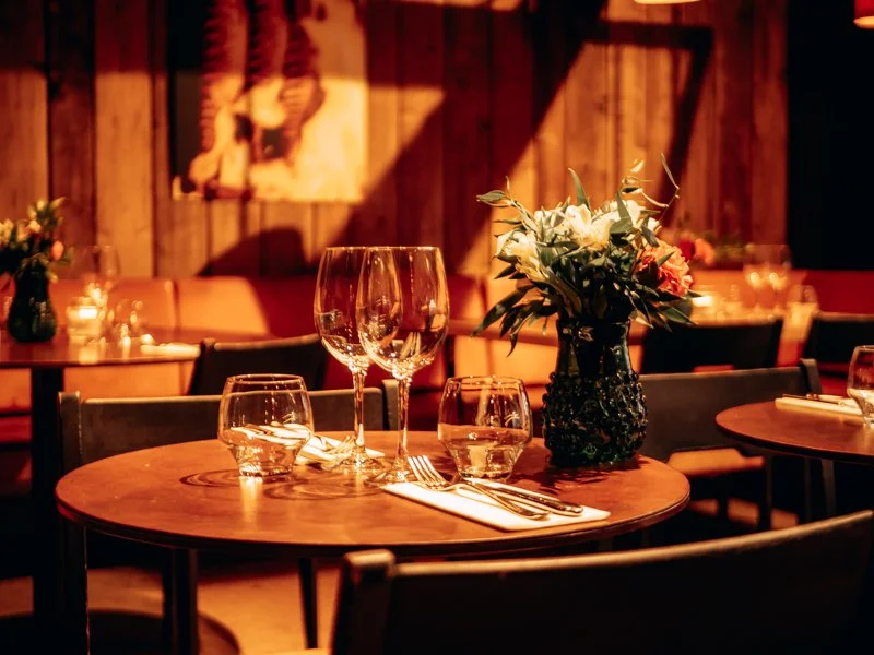 A table set for dining with wine glasses, water glasses, a floral centerpiece, and silverware, in a dimly lit restaurant with wooden walls and warm lighting.