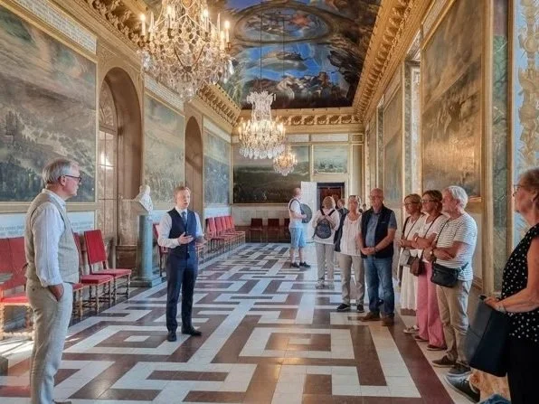 Tour guide speaking to visitors in a luxurious room with ornate chandeliers, marble floors, and classical paintings on the walls.