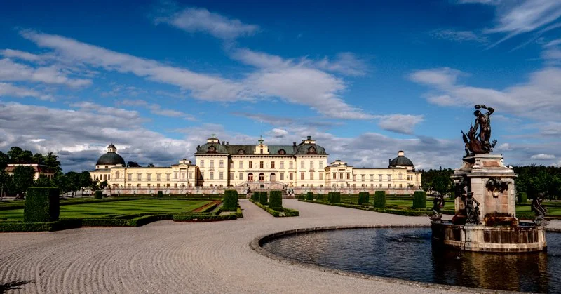A large historic palace with a symmetrical design, surrounded by manicured gardens and pathways, with a fountain and statue in the foreground, under a partly cloudy sky.