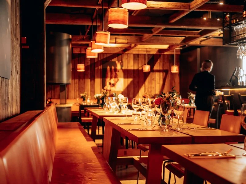 A warmly lit restaurant with wooden decor, tables set with wine glasses, flowers, and cutlery, and a server in the background.