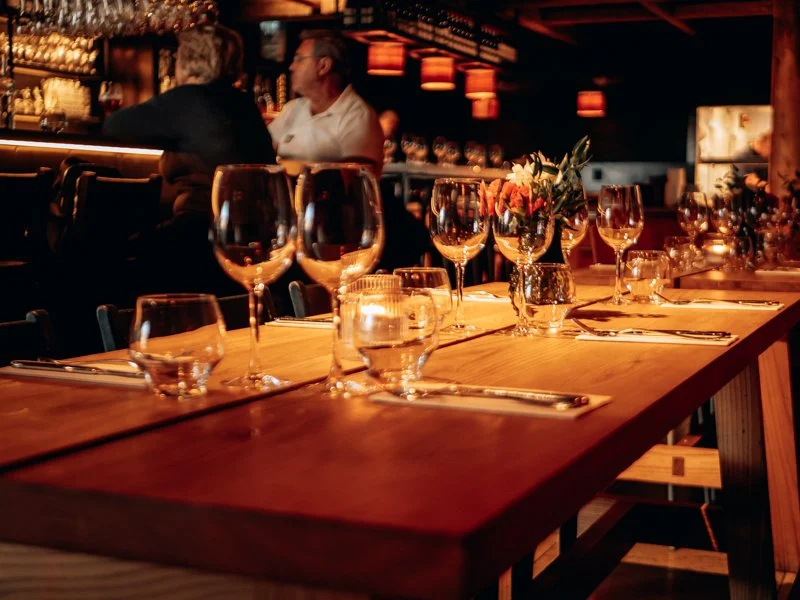 A table set for dining with wine glasses, water glasses, silverware, and a floral centerpiece in a dimly lit restaurant.