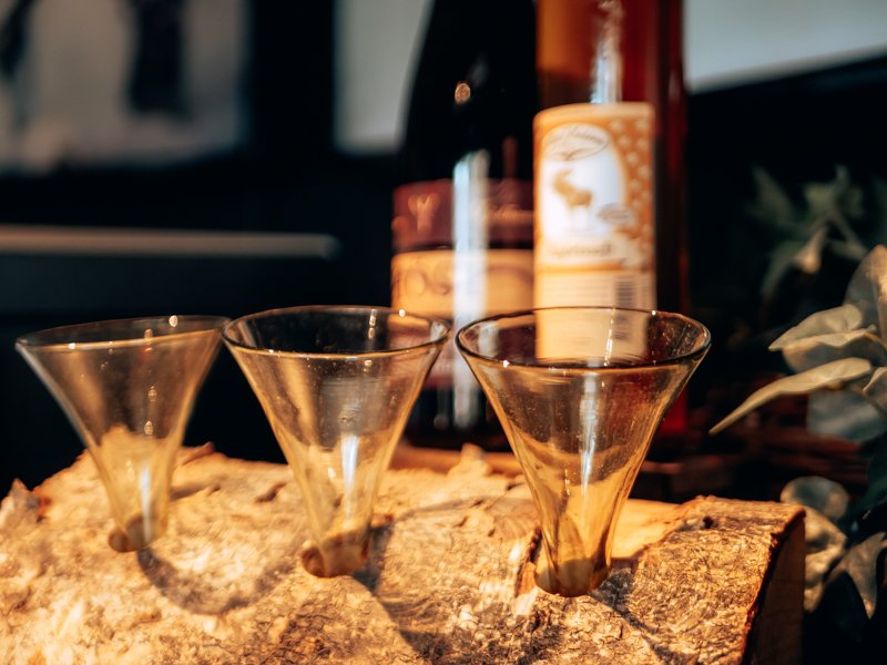 Three empty shot glasses on a wooden surface with bottles of alcohol in the background.