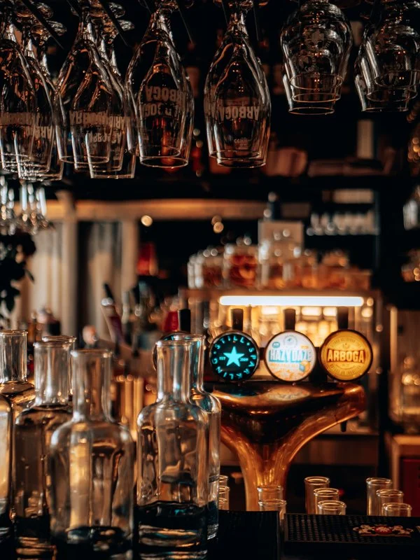 Bar with hanging wine glasses, empty glass bottles, and beer tap handles, illuminated by warm lighting.