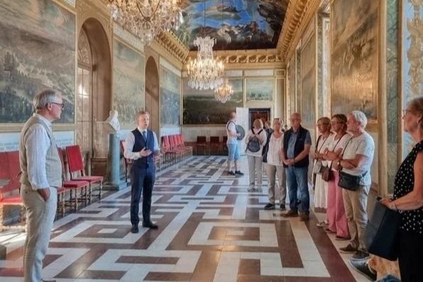 A tour guide in a blue vest and white shirt explaining to a group of tourists in an ornate, decorated room with chandeliers, large artworks on the walls, and a patterned floor.