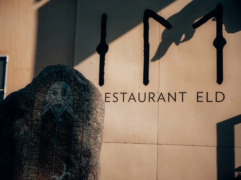 Shadow of a hand holding an umbrella over a wall with the text 'RESTAURANT ELD' and a stone with a carved face.