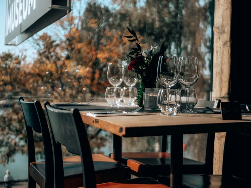 A table set for a meal outdoors with empty wine glasses, water glasses, silverware, and a floral centerpiece, with fall foliage visible in the background.