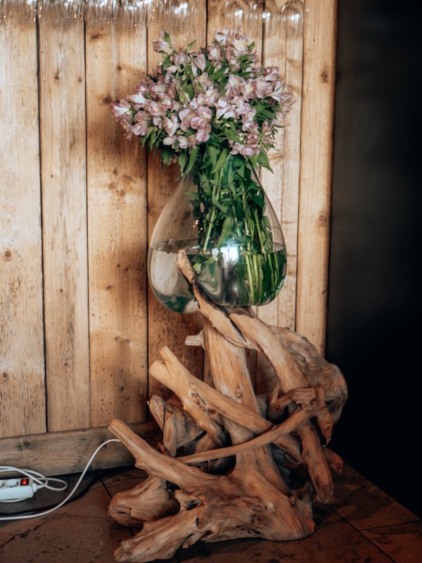 A large glass vase with pink and white flowers on a wooden driftwood stand against a wooden wall.