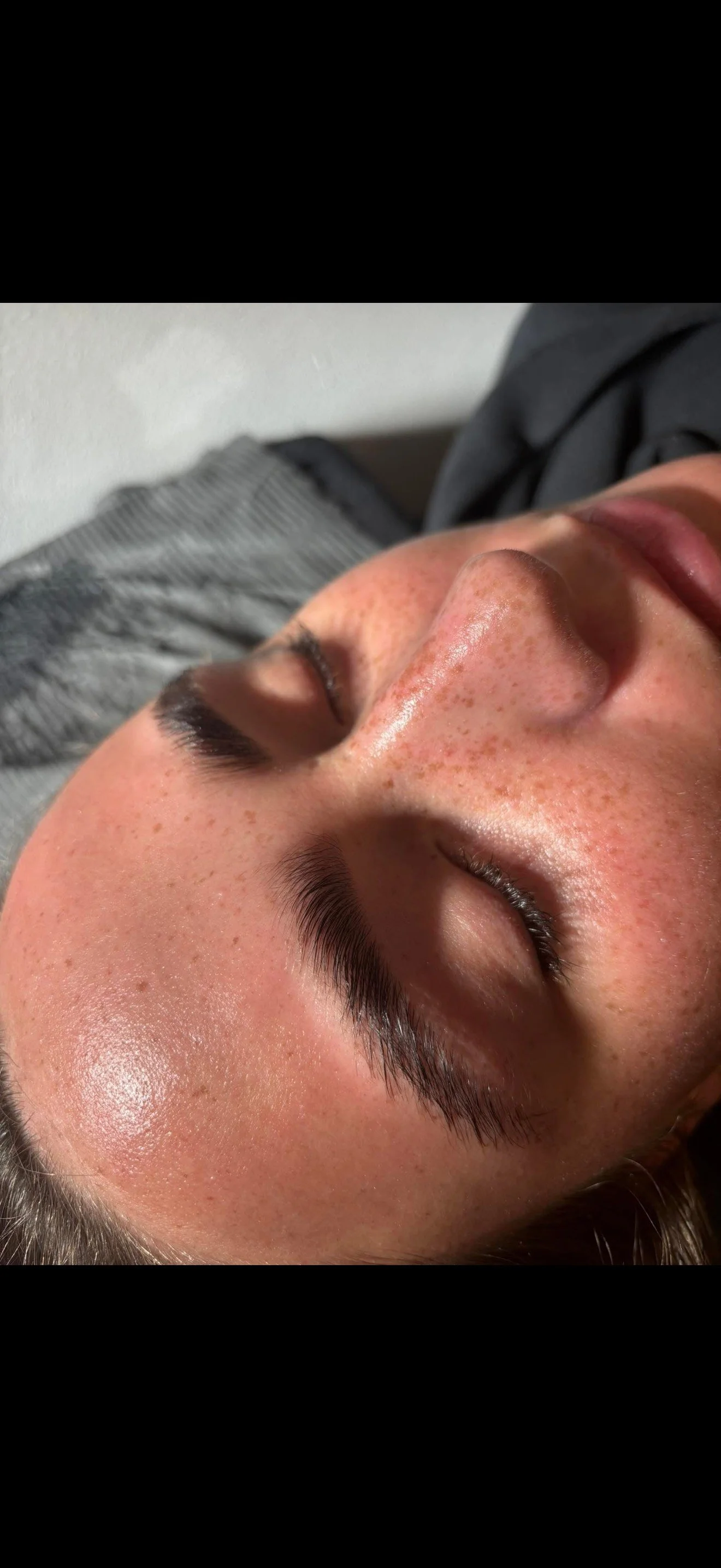 Close-up of a person's face with closed eyes, light skin with freckles, natural eyebrows, and shiny skin. The person appears relaxed, lying on a bed or couch.