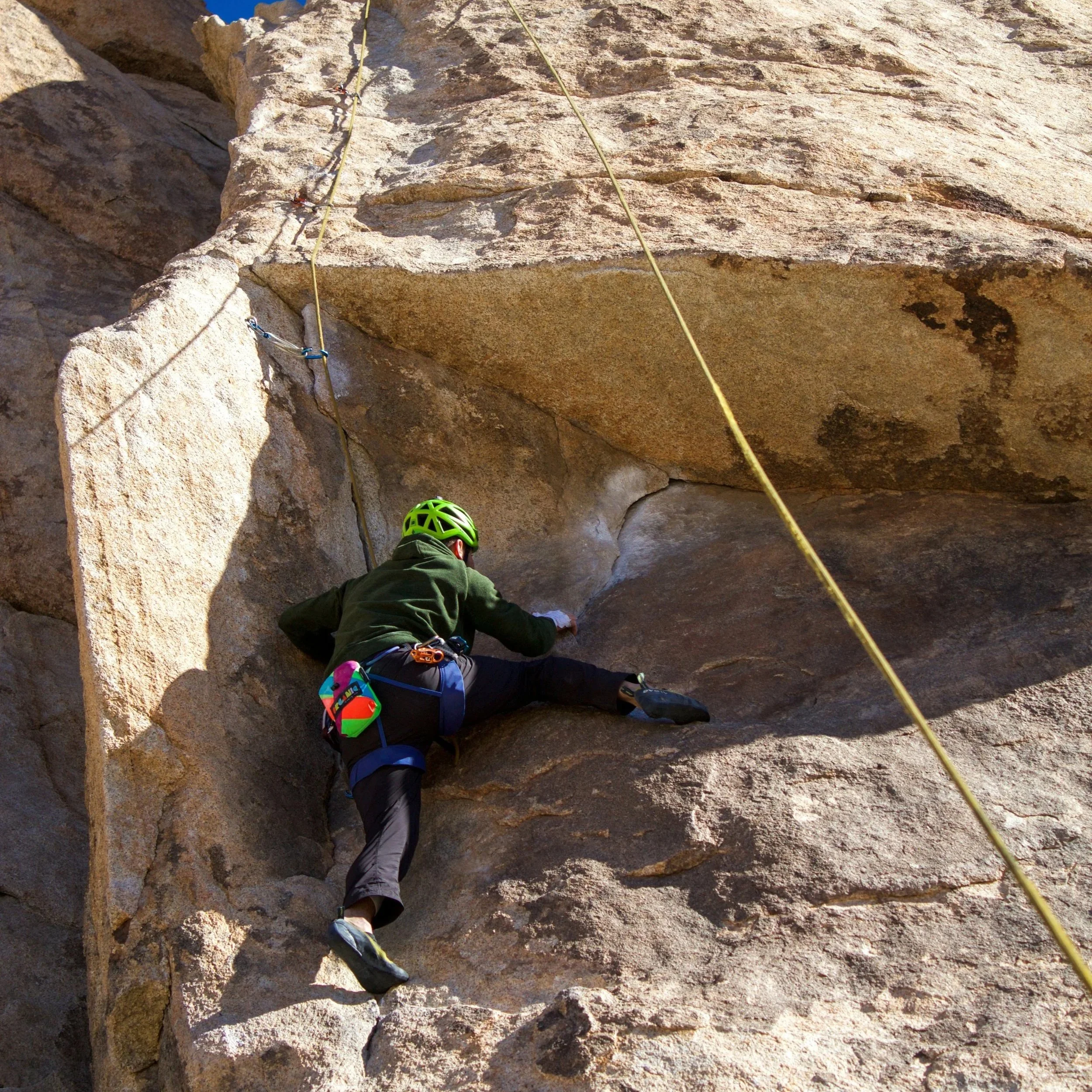 A person wearing a green helmet and climbing gear is rock climbing on a steep rock face with a rope attached for safety.