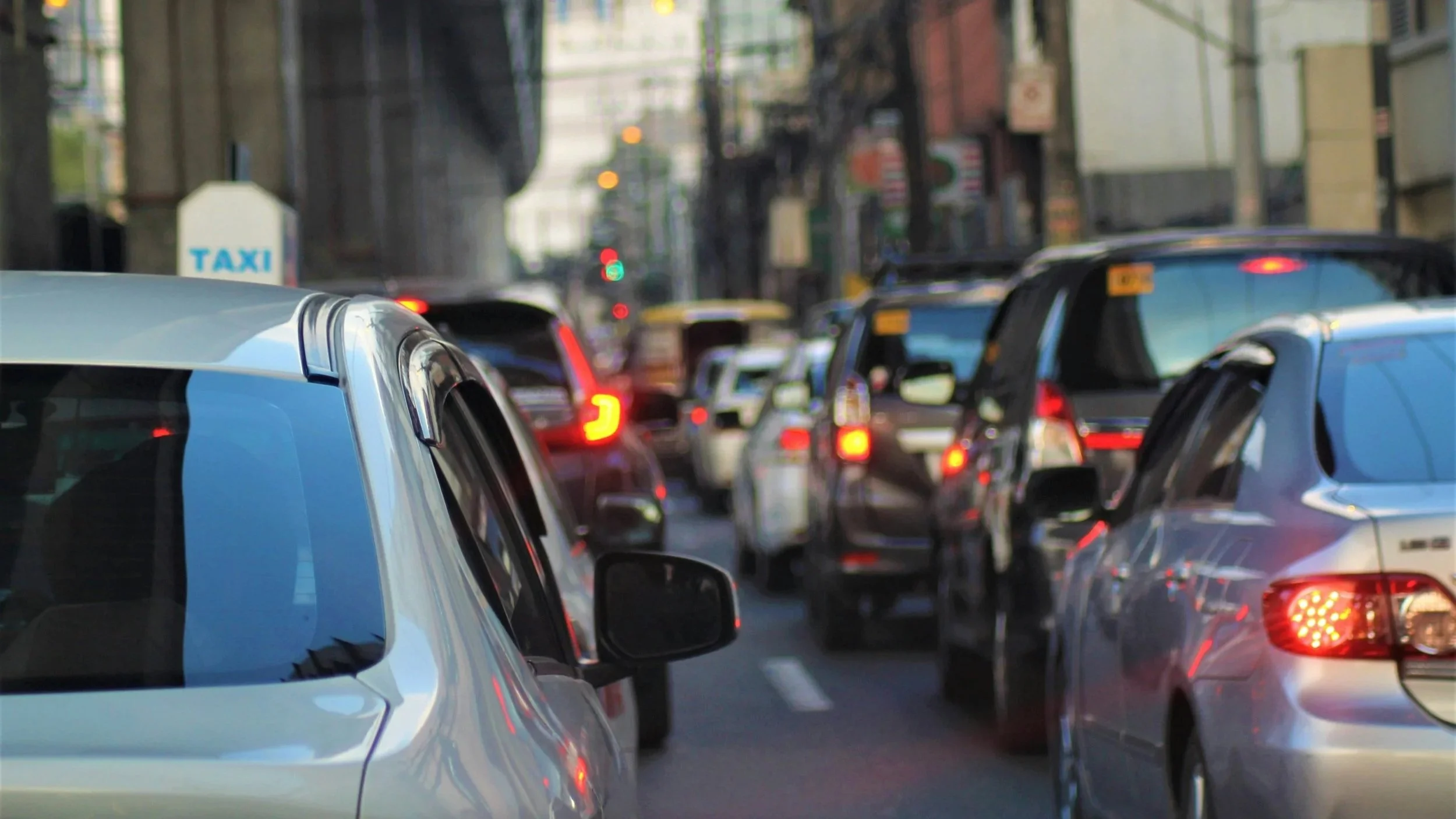 Traffic jam with multiple cars lined up on city street, red brake lights illuminated, tall buildings on either side, and a taxi sign visible in the background.