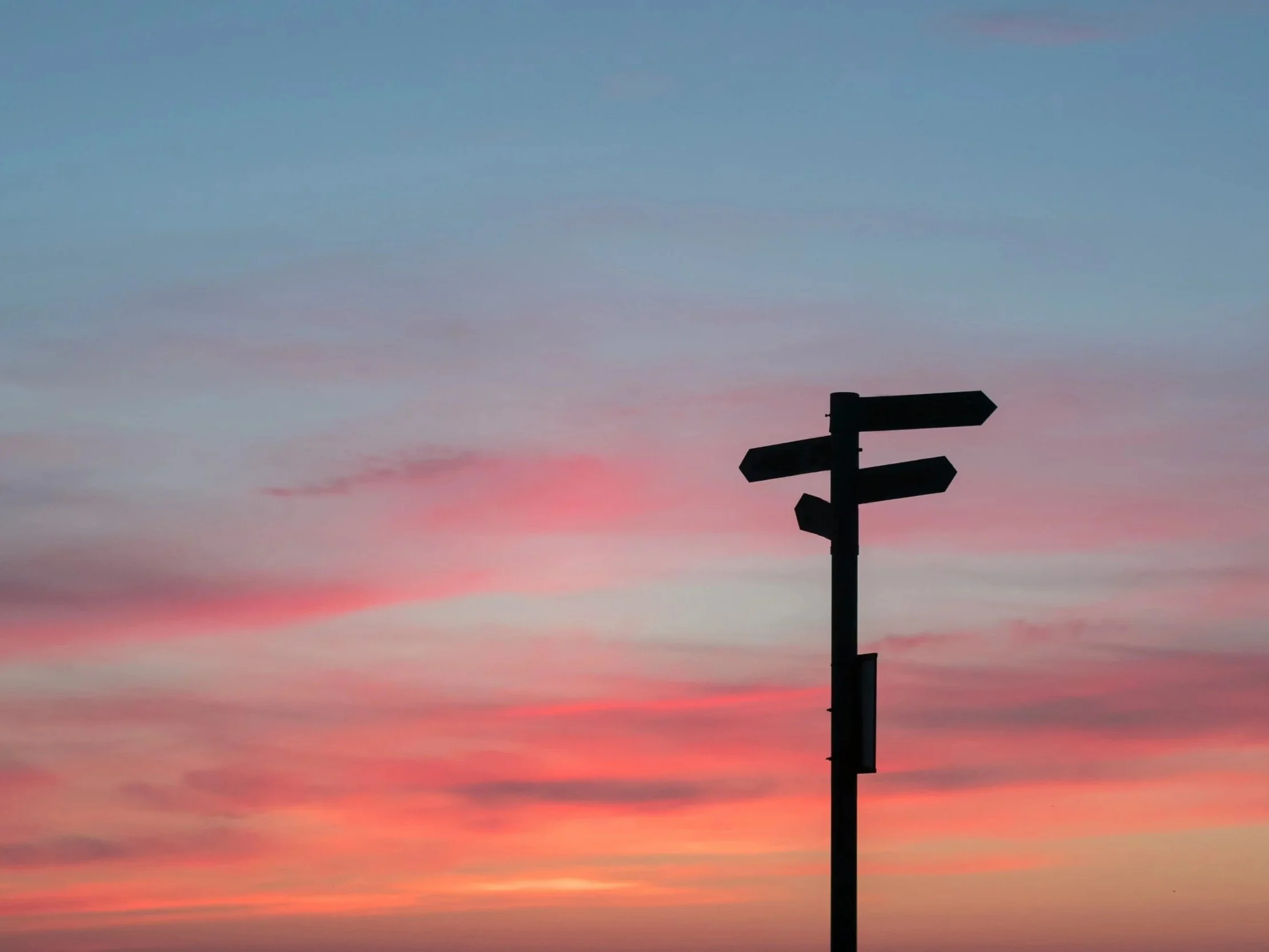 Silhouette of a multi-directional signpost against a colorful sunset sky with pink, orange, and blue clouds.