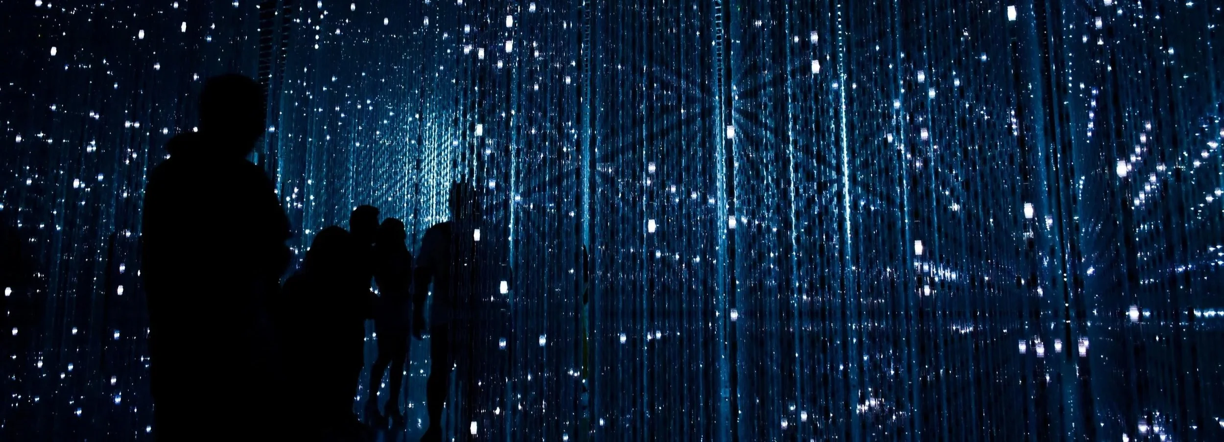 People standing inside a dark room filled with hanging blue lights, creating an immersive light installation.