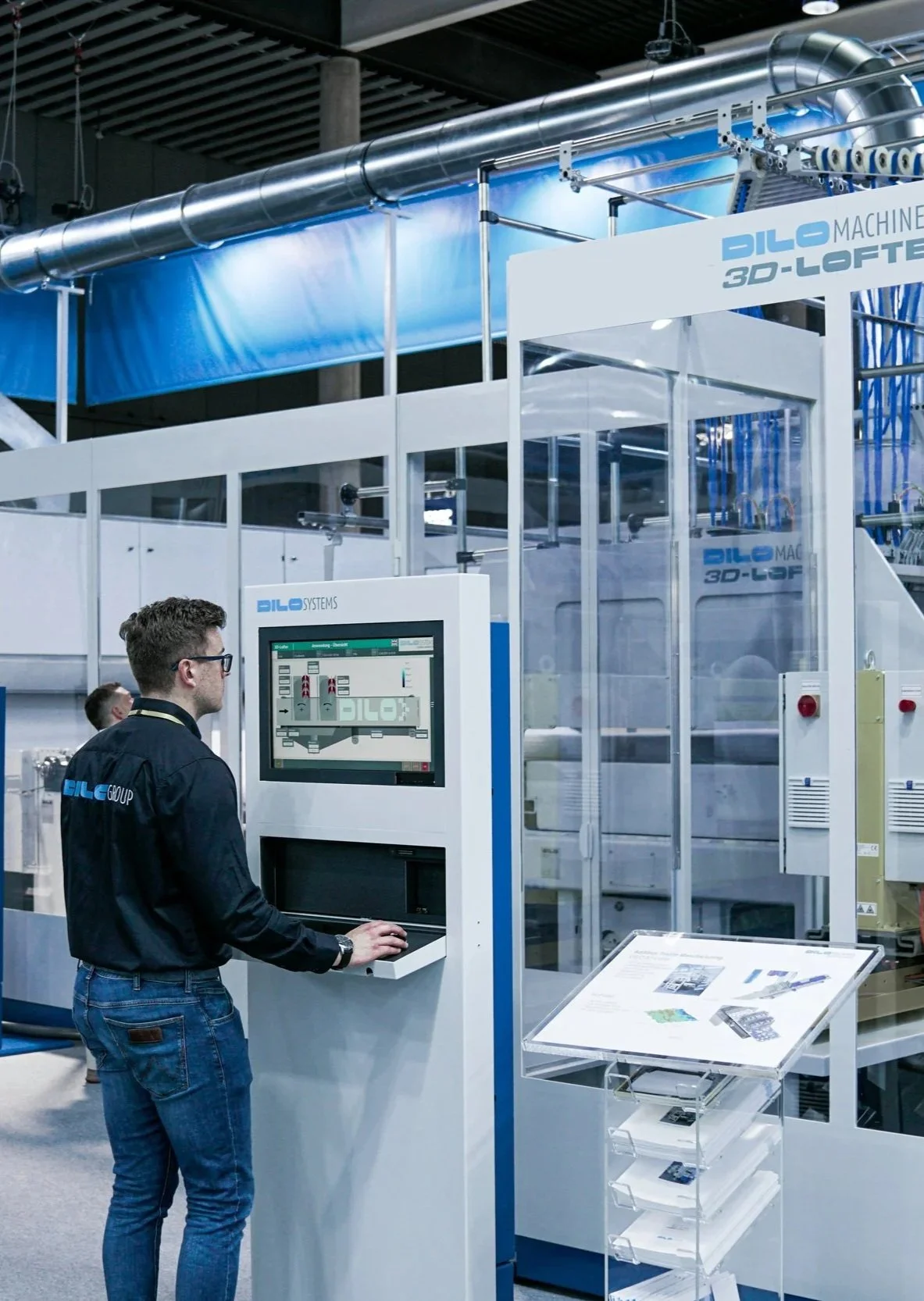 A man operating a control panel at a machinery exhibit, with a large industrial machine labeled 'DILO 3D-LOFT' and 'DILO SYSTEMS' in the background.