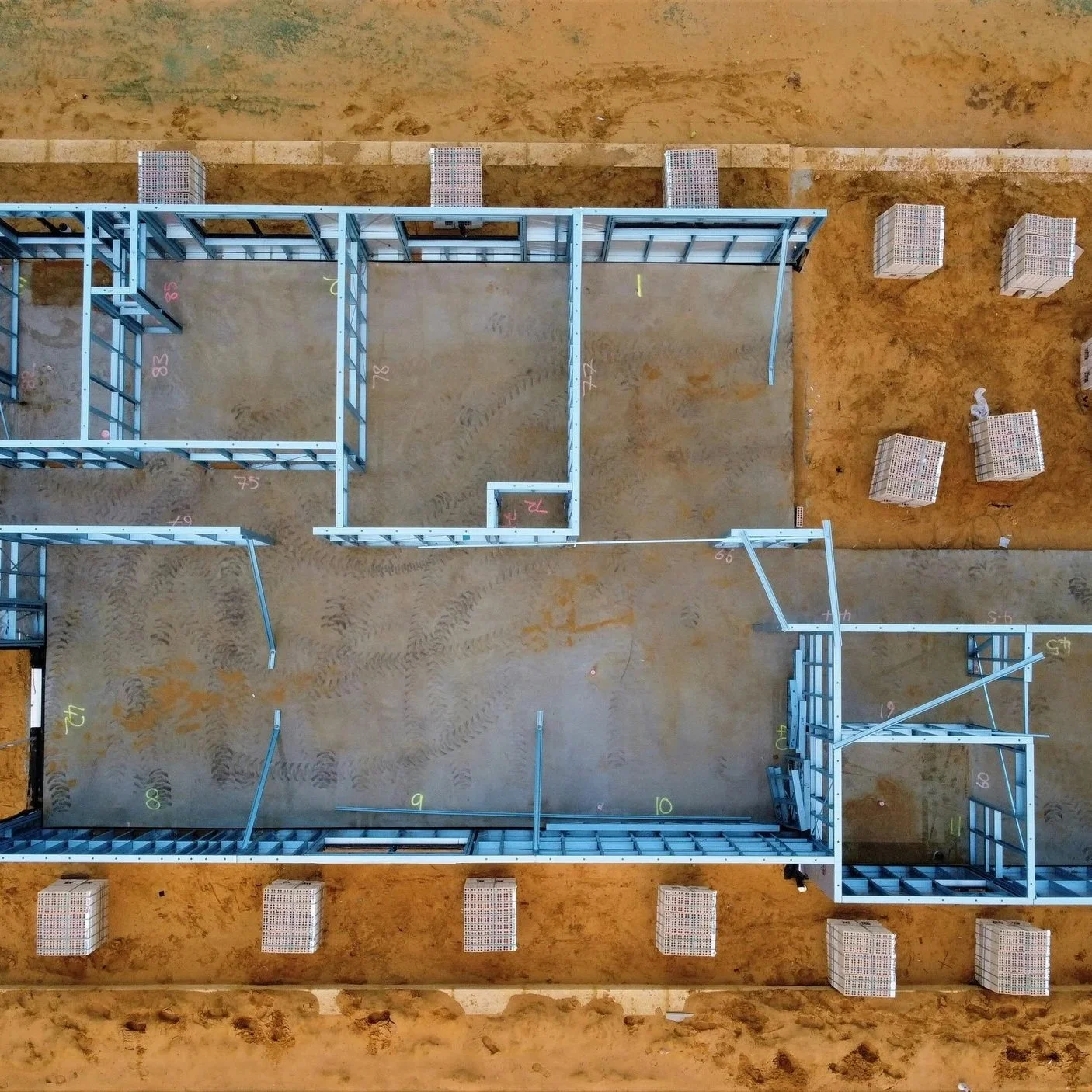 An overhead view of a construction site showing the metal framing for a building with numbered markings. Surrounding the framing, there are several stacks of materials and footprints in the dirt.