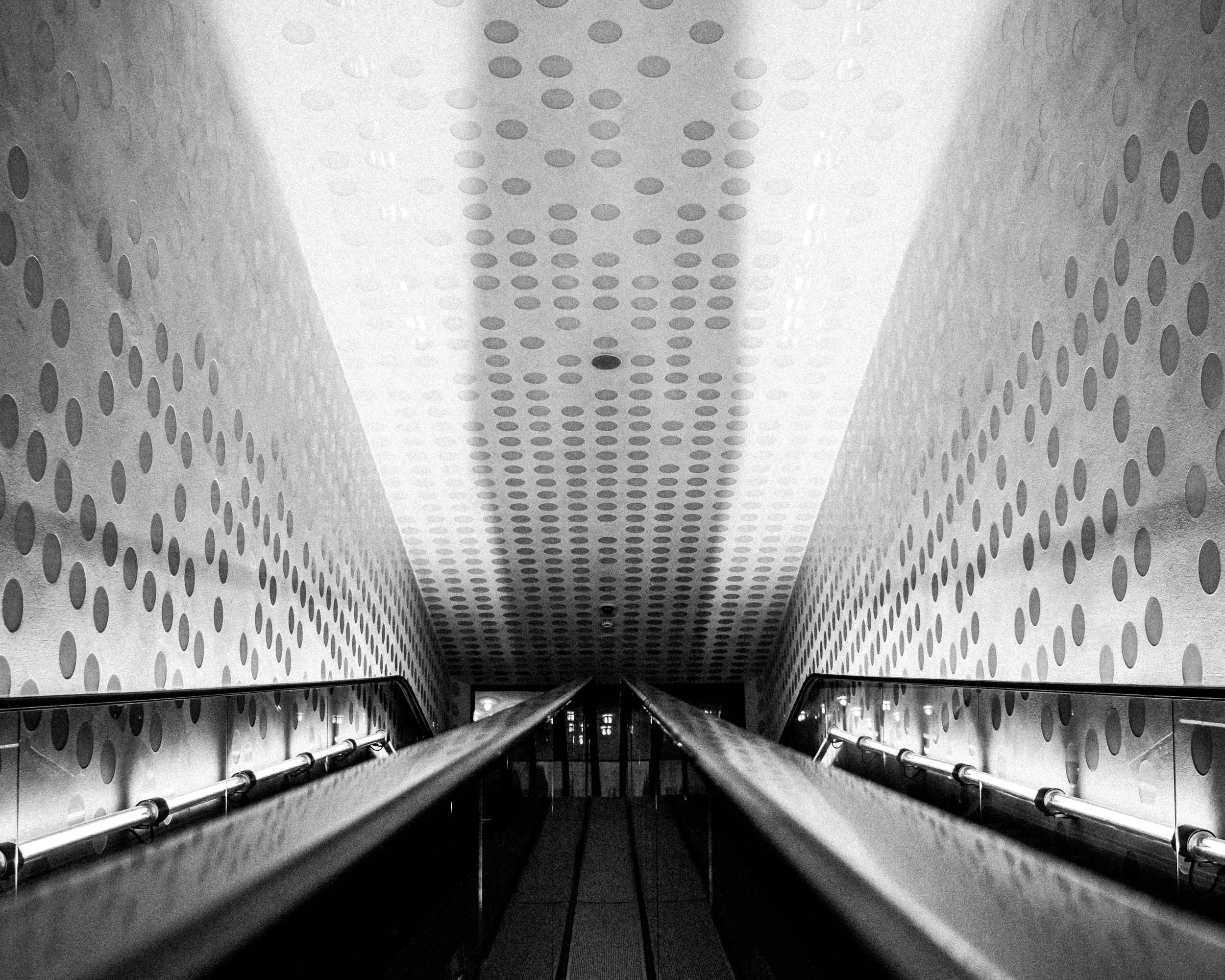 Black and white photo of a downward view of an escalator at a modern building with perforated wall panels.