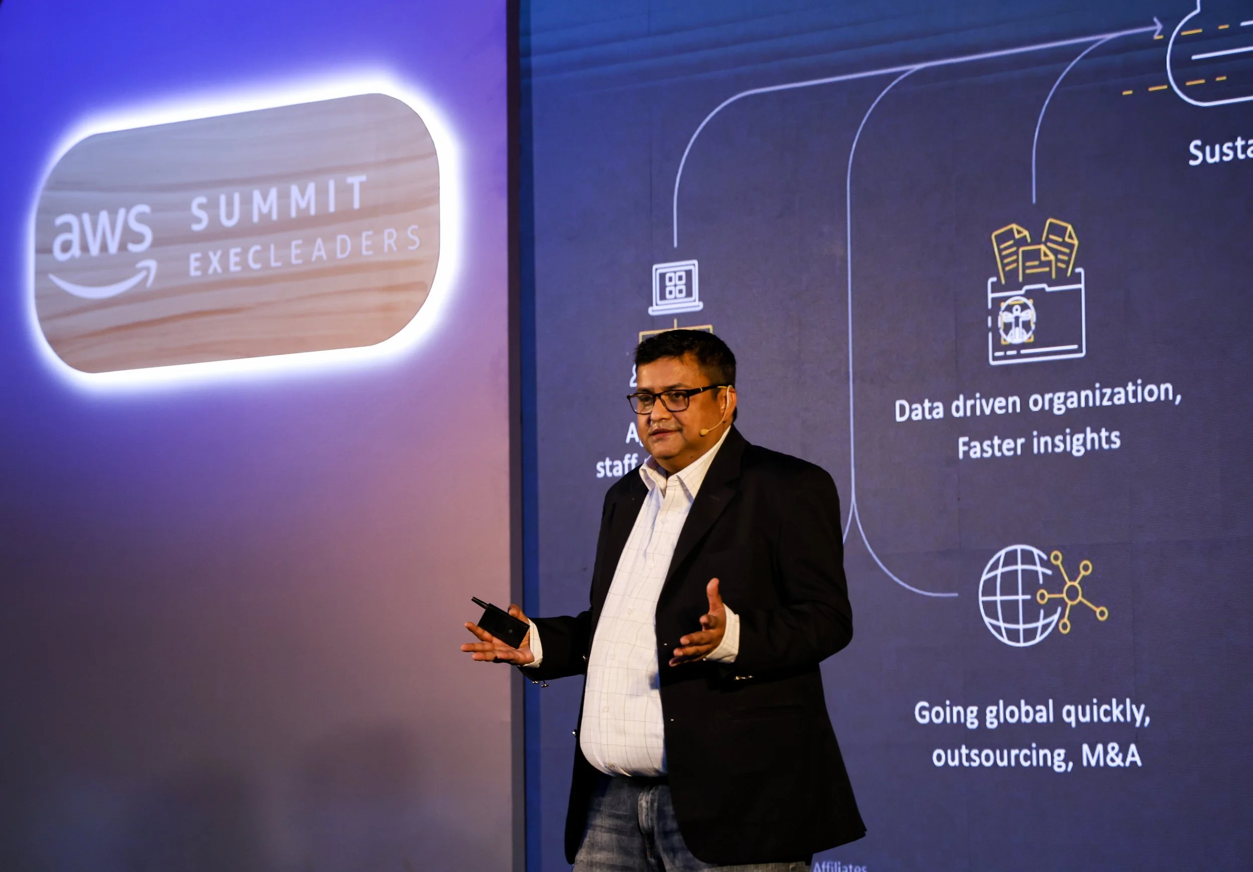 A man in a black blazer and white shirt giving a presentation at an AWS Summit Excelerators event. The presentation slide shows icons and text about data-driven organization and going global quickly.
