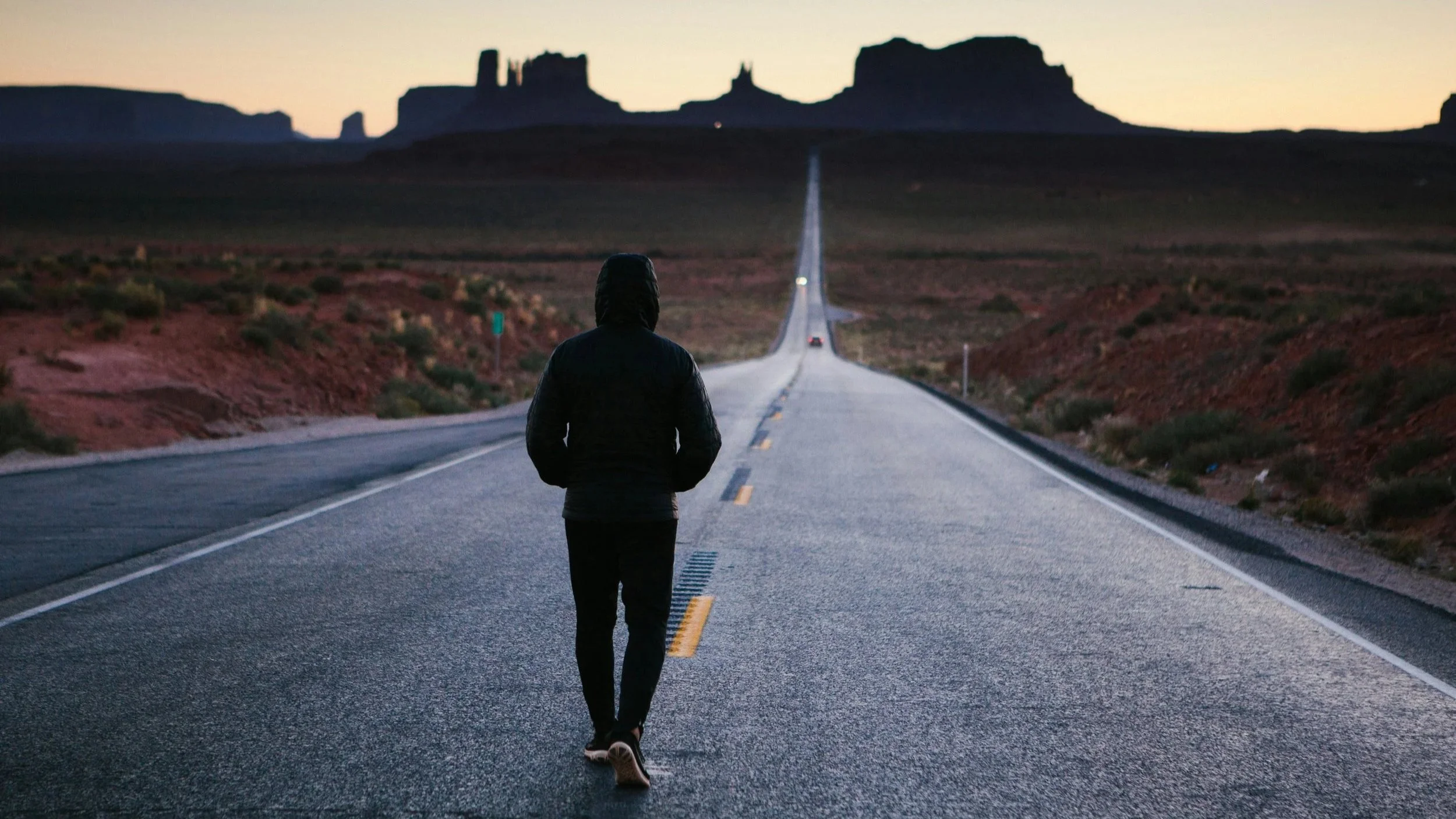 Person walking alone on an empty highway in desert landscape during sunset or dusk with rock formations in the background.